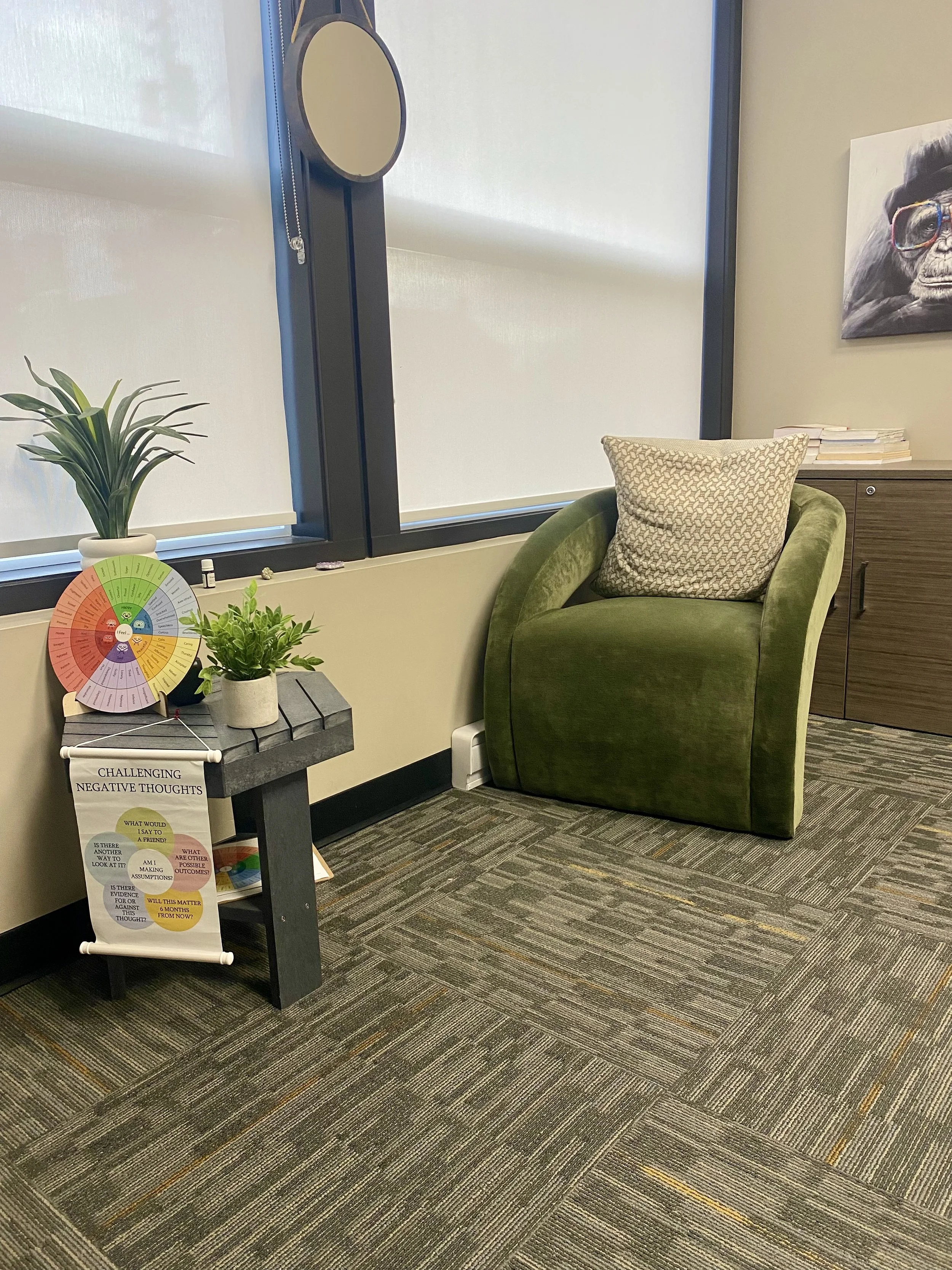 An office space with a green armchair and a patterned pillow. A small table with plants and educational materials, a window with shades, a wooden cabinet with books, and wall art including a gorilla wearing glasses.
