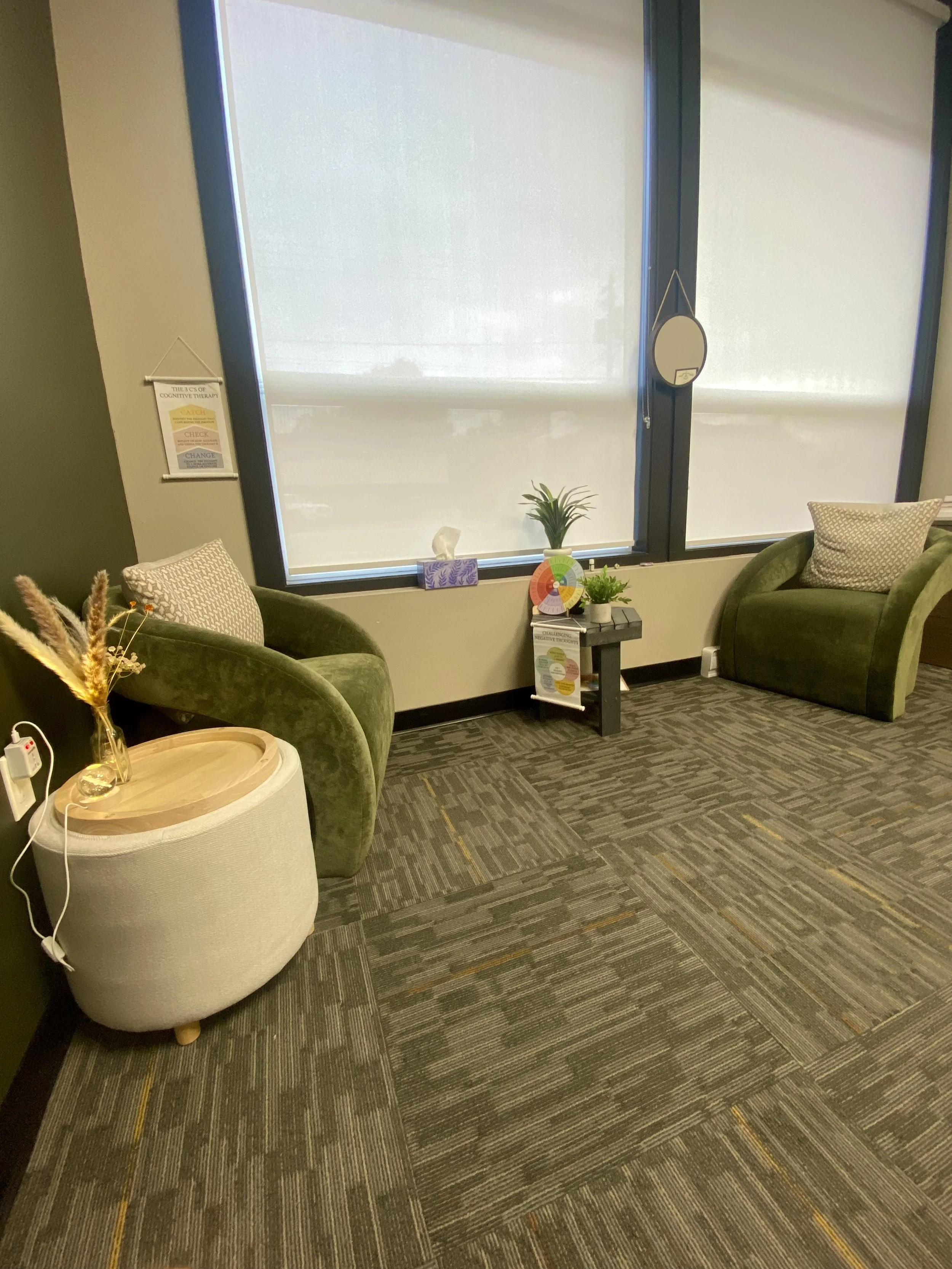 A waiting room with two green velvet armchairs with patterned pillows, a small side table with plants and a colorful wheel, a large window with closed blinds, and a framed motivational poster on the wall.