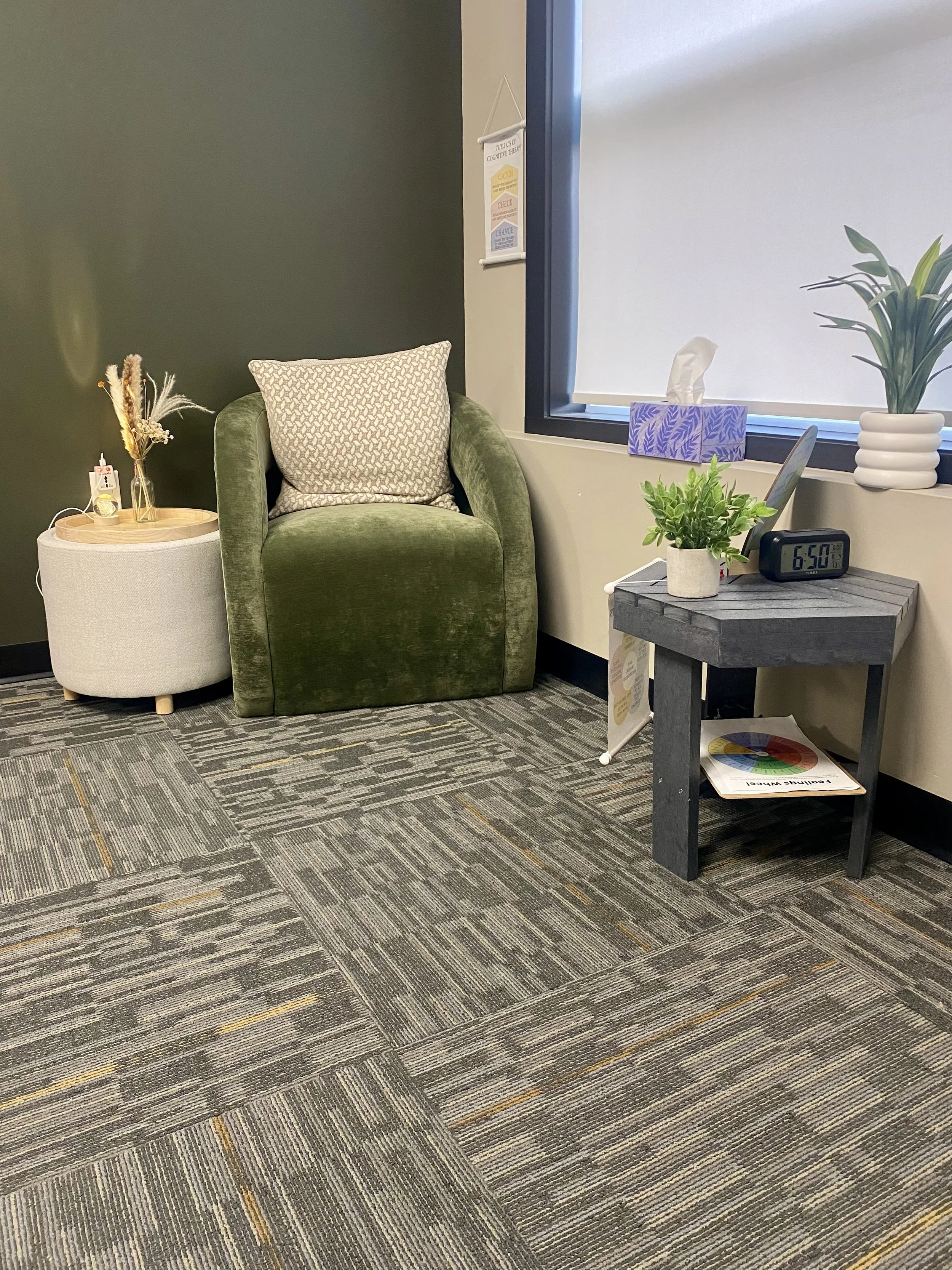 A cozy waiting area with an olive green armchair, a small round white side table with decorative dried plants, and a gray wooden table with a potted plant, a digital clock showing 6:50, and a box of tissues near a window with a roller blind.