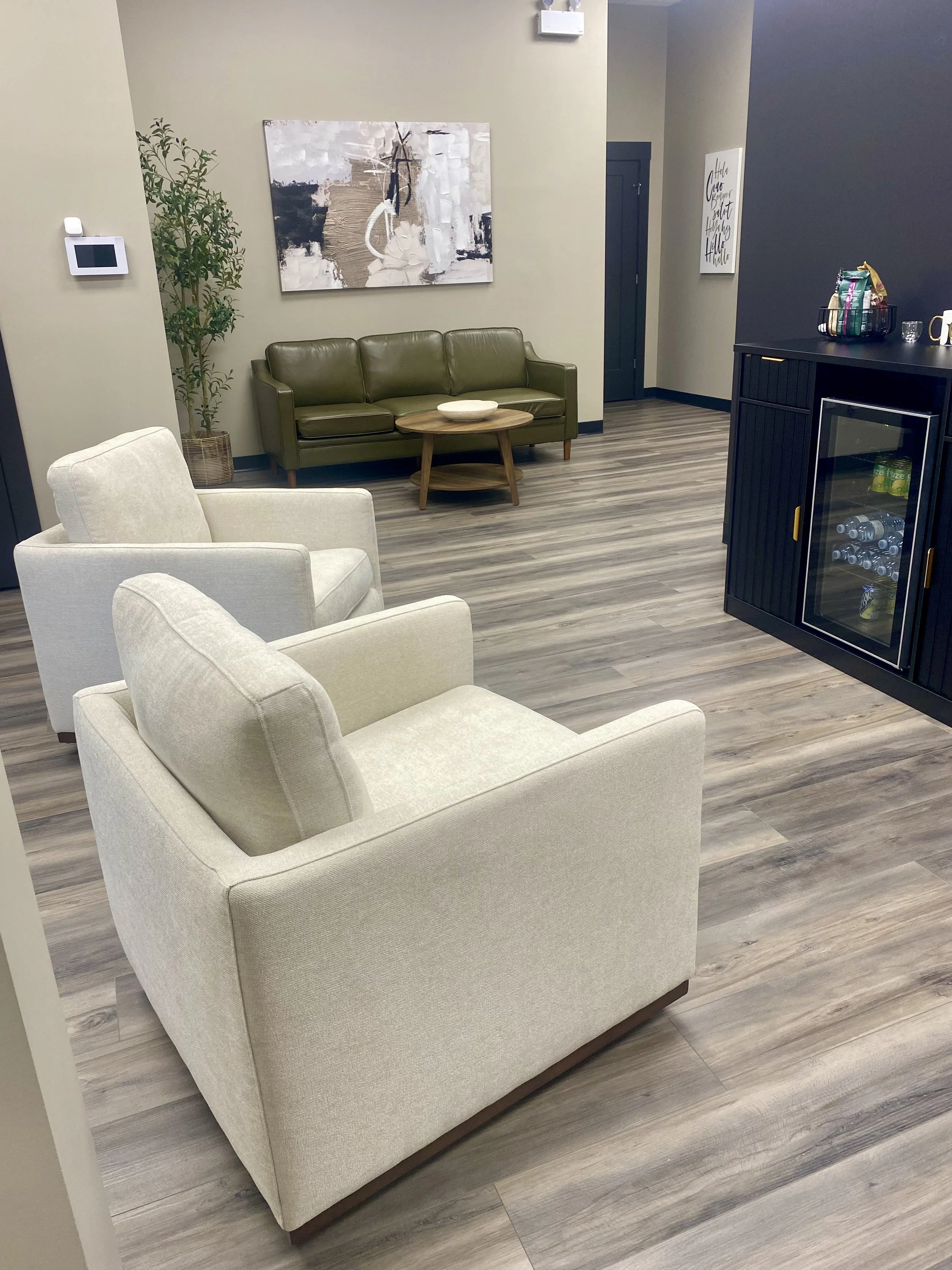 A contemporary living room with cream upholstered armchairs, a green leather sofa, a round wooden coffee table, a tall potted plant, abstract wall art, and a black cabinet with a mini fridge and snacks on top.