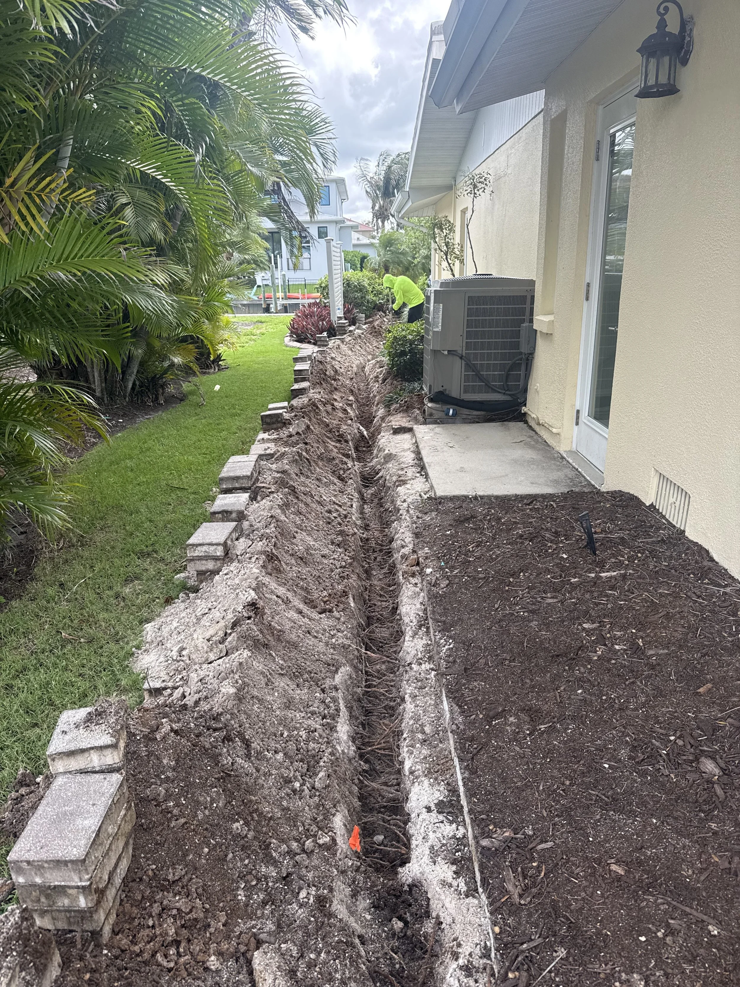 A backyard with a trench dug parallel to the house, with concrete blocks positioned along the edge of the trench. A worker in a neon green shirt is digging further down the trench. The yard has green grass, bushes, and palm trees, with some houses and a pool visible in the background.