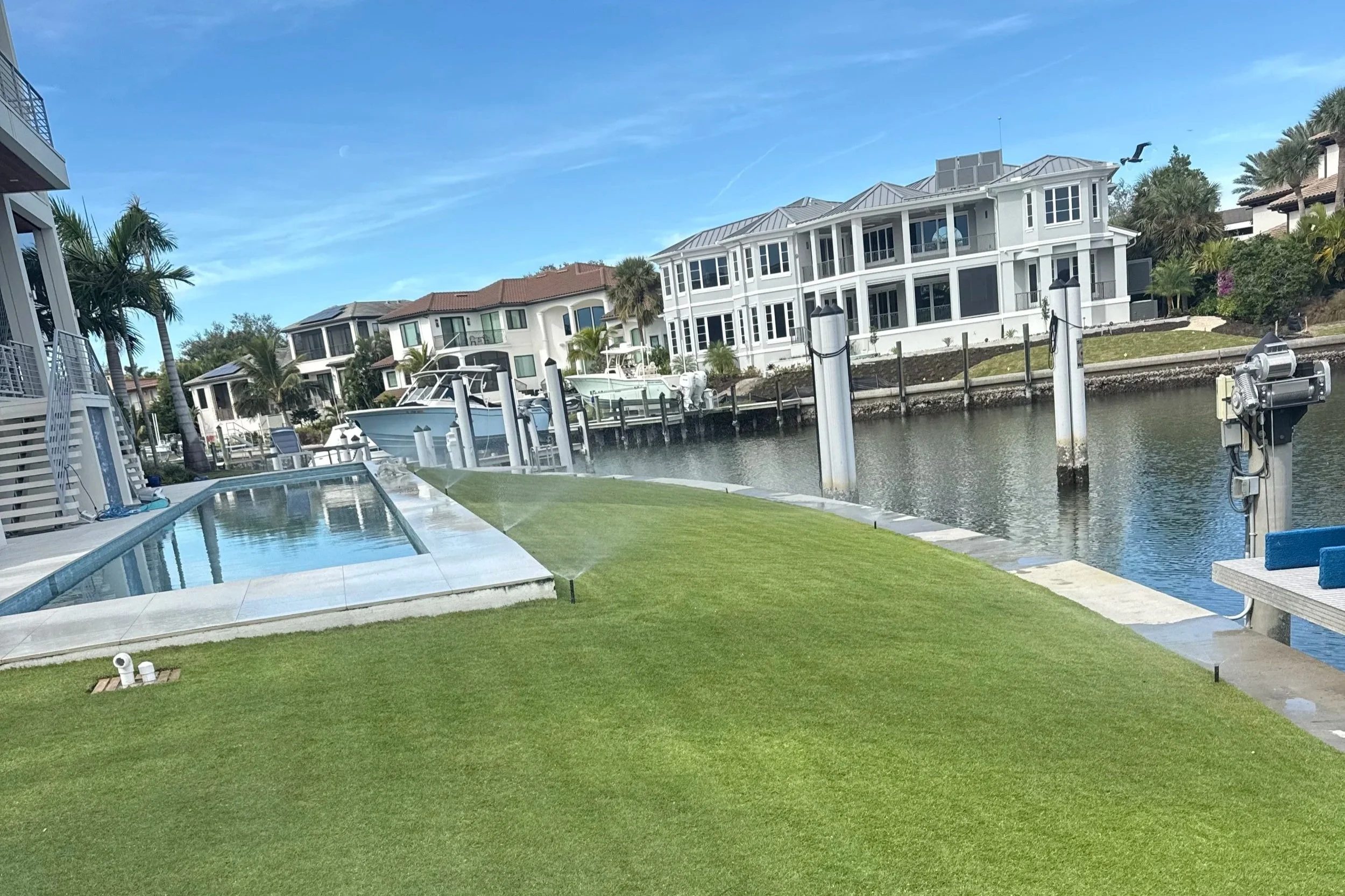A waterfront residential area with modern houses, boats docked at a marina, a swimming pool, and lush green lawn under a blue sky.