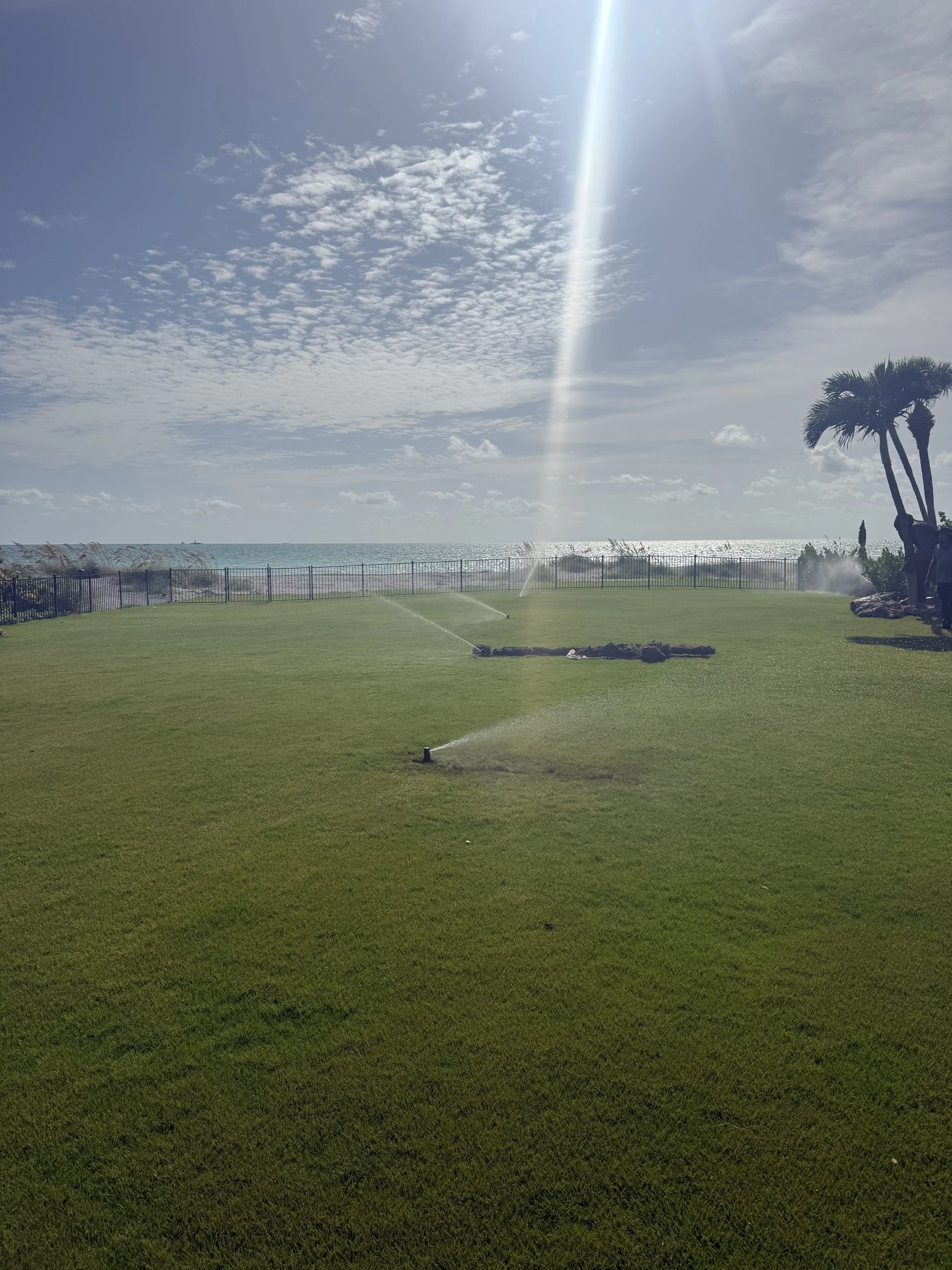 A green grassy area with sprinkler systems watering the lawn. In the background, there is a fence, palm trees, and the ocean with ships in the distance. The sky is partly cloudy with a visible beam of sunlight.