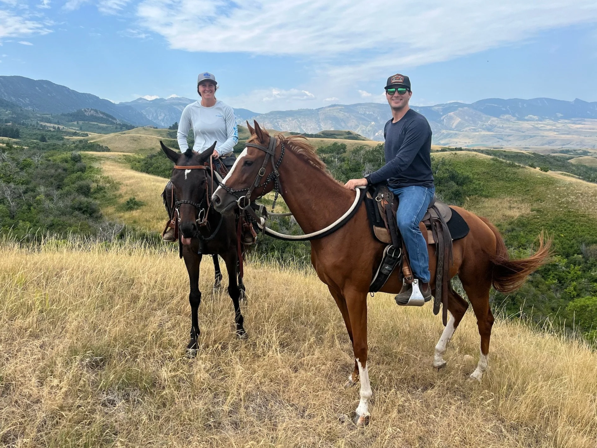 Two people riding horses in a grassy field with rolling hills and mountains in the background. The woman on the left is wearing a gray cap and white long-sleeve shirt, and the man on the right is wearing a black cap, sunglasses, and a dark long-sleeve shirt.