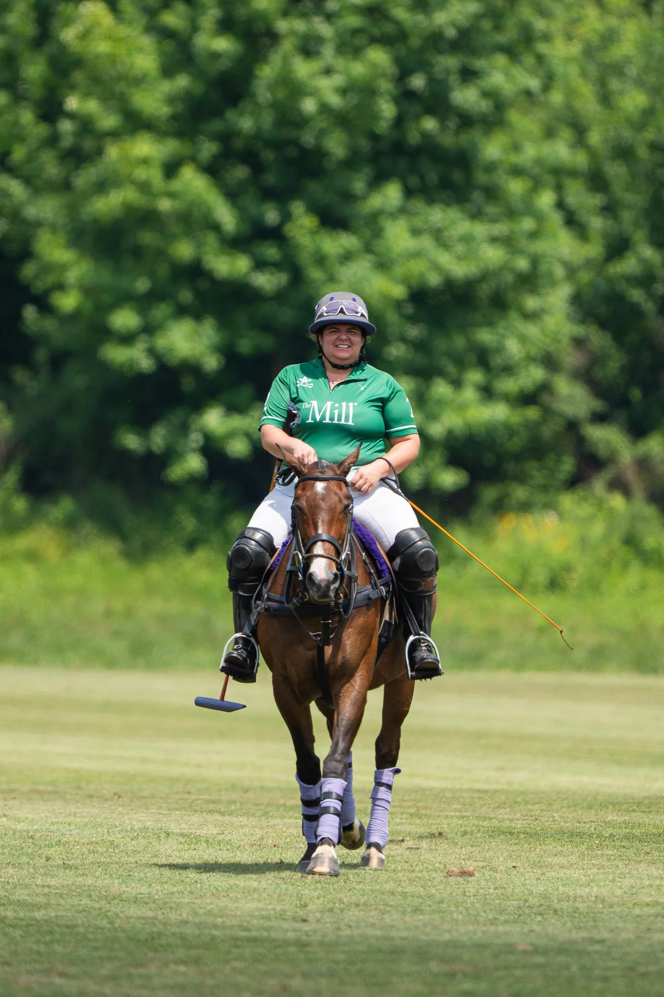 A woman riding a polo horse on a green field during daytime. She is wearing a green shirt, riding gear, and a helmet with goggles. The horse is equipped with polo gear, including leg protection, and they are moving towards the camera with trees in th
