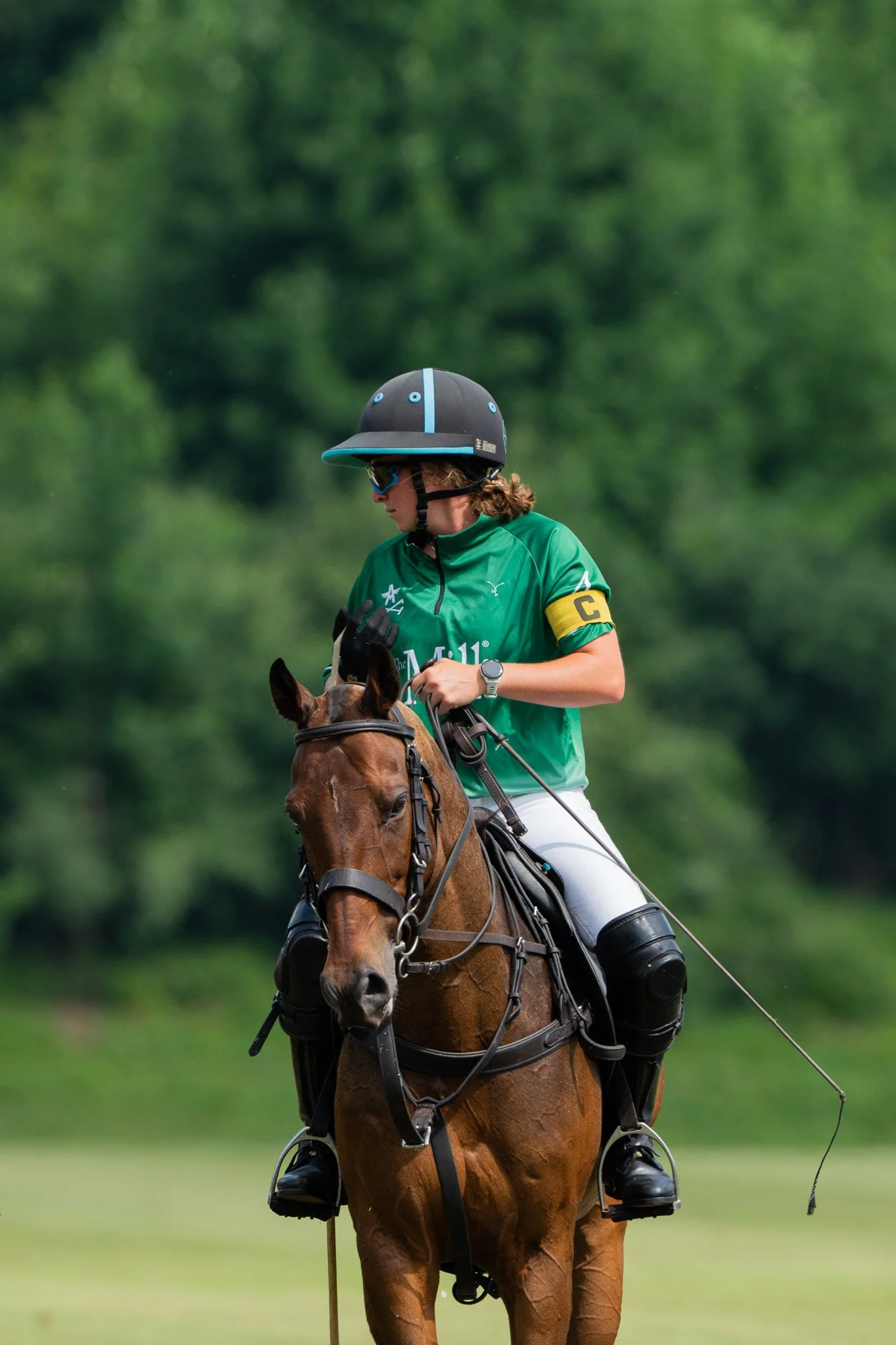 A female polo player wearing a green jersey with a yellow captain's armband and a black helmet, riding a brown horse on a field with green trees in the background.