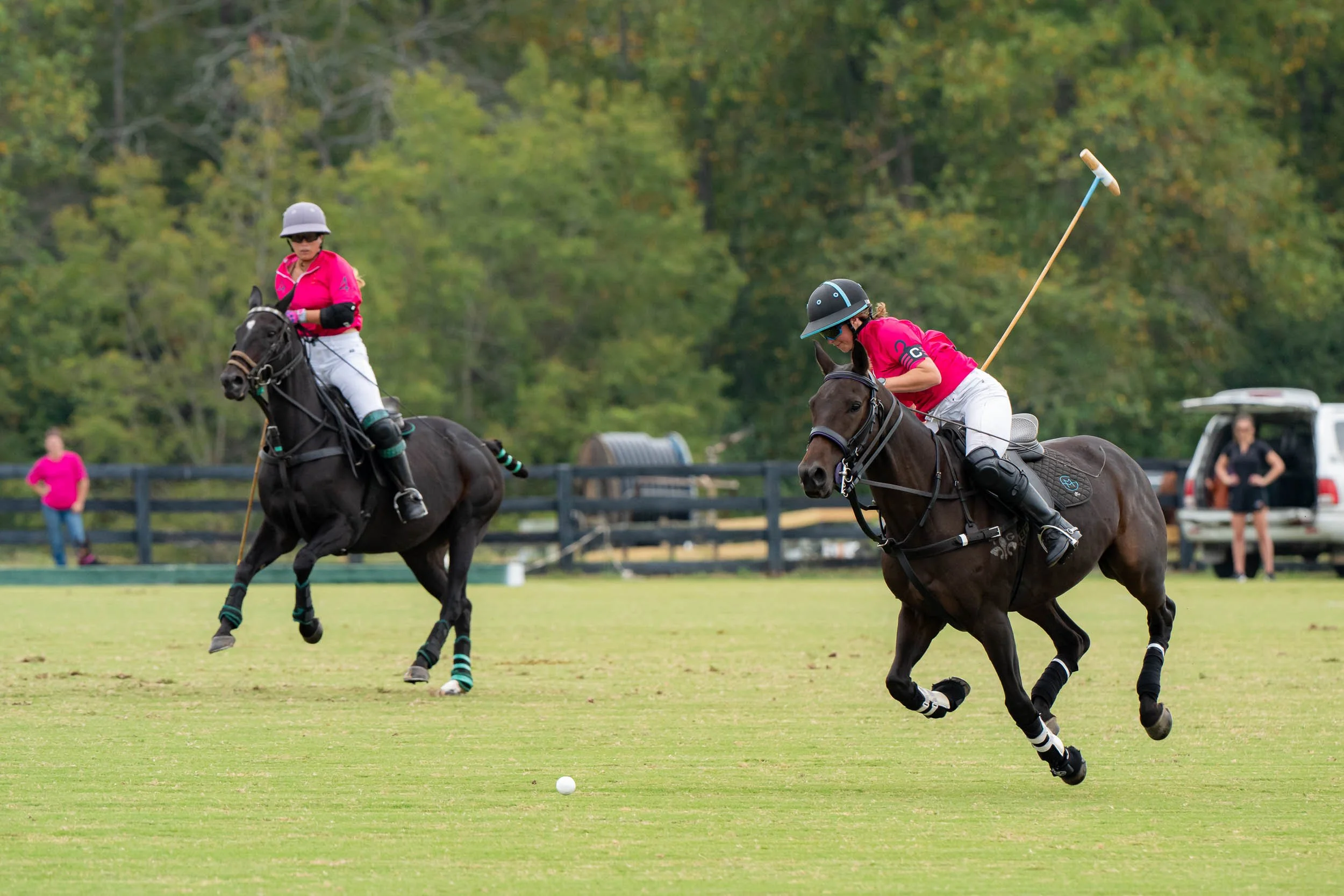 Polo players in pink jerseys riding horses and chasing a ball on a grassy field.
