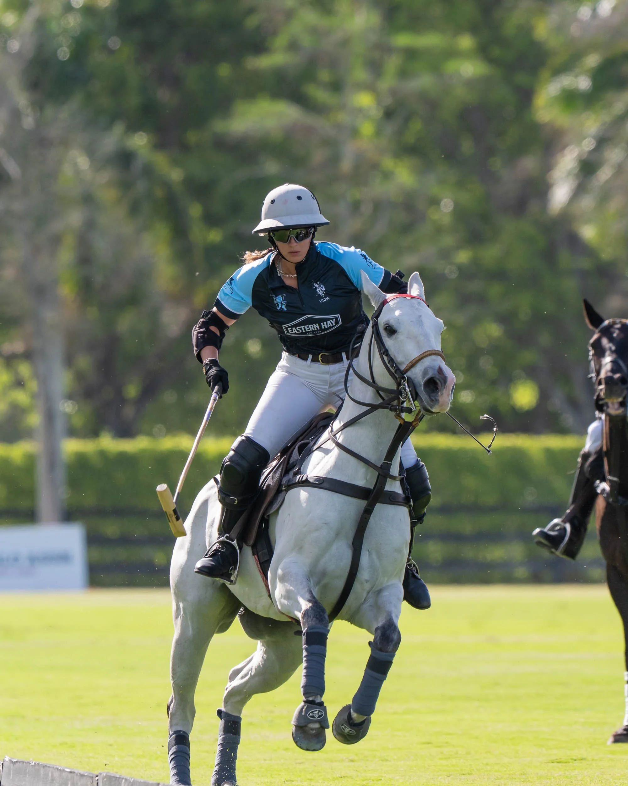 Female polo player wearing protective helmet and sunglasses riding white horse during match on green field with trees in background.