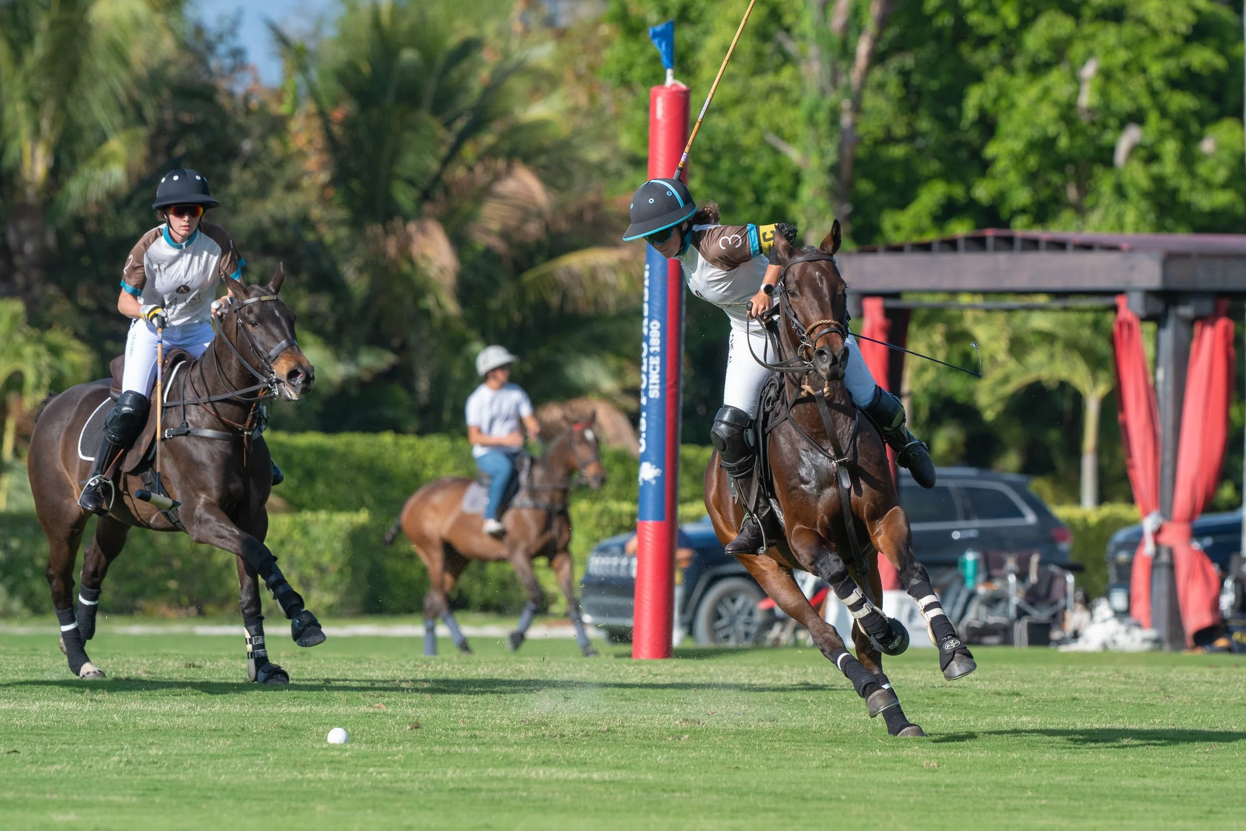 Two polo players on horseback competing for the ball during a polo match, with a third player and spectators in the background.