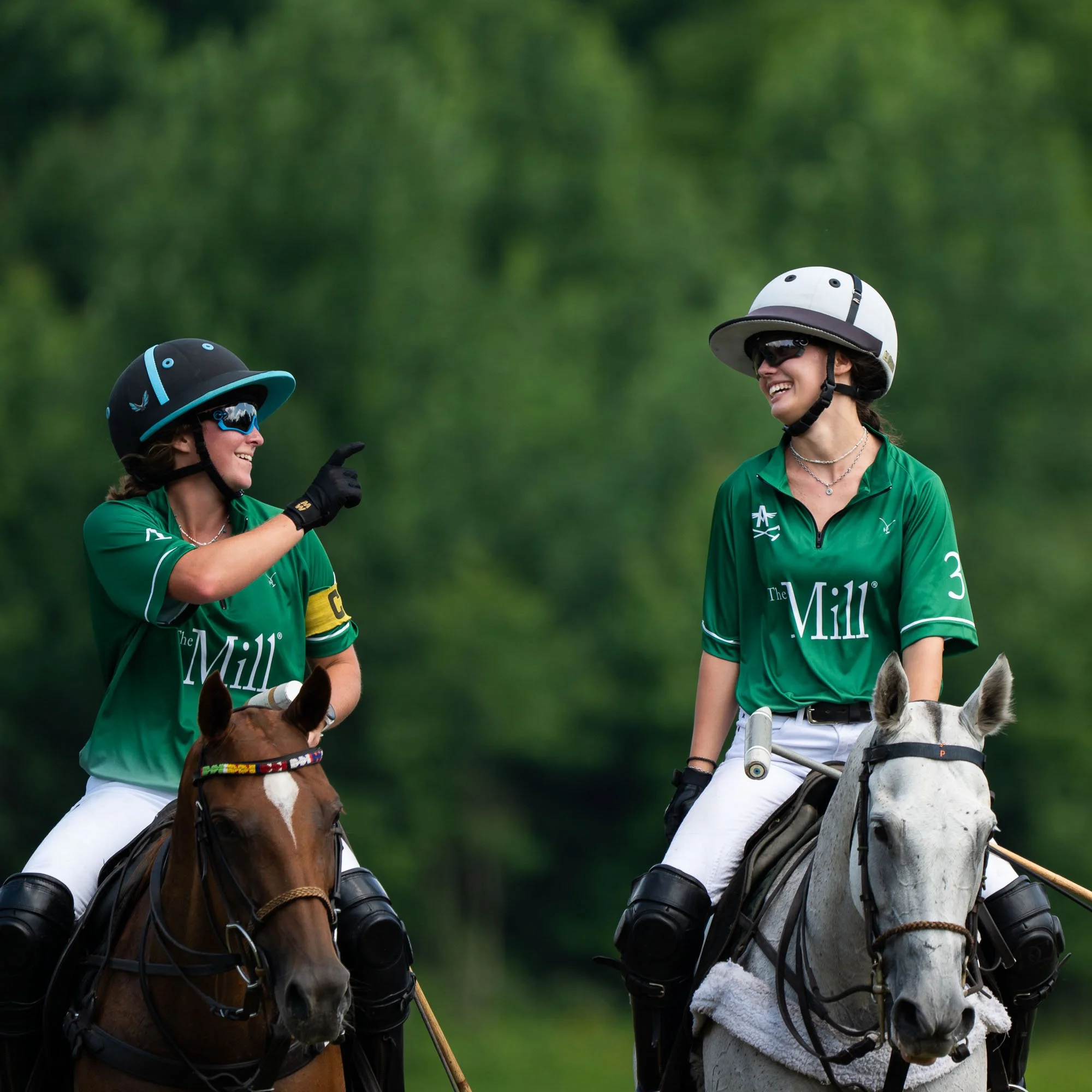 Two women wearing green shirts, helmets, and sunglasses riding horses and smiling at each other against a blurred green background.