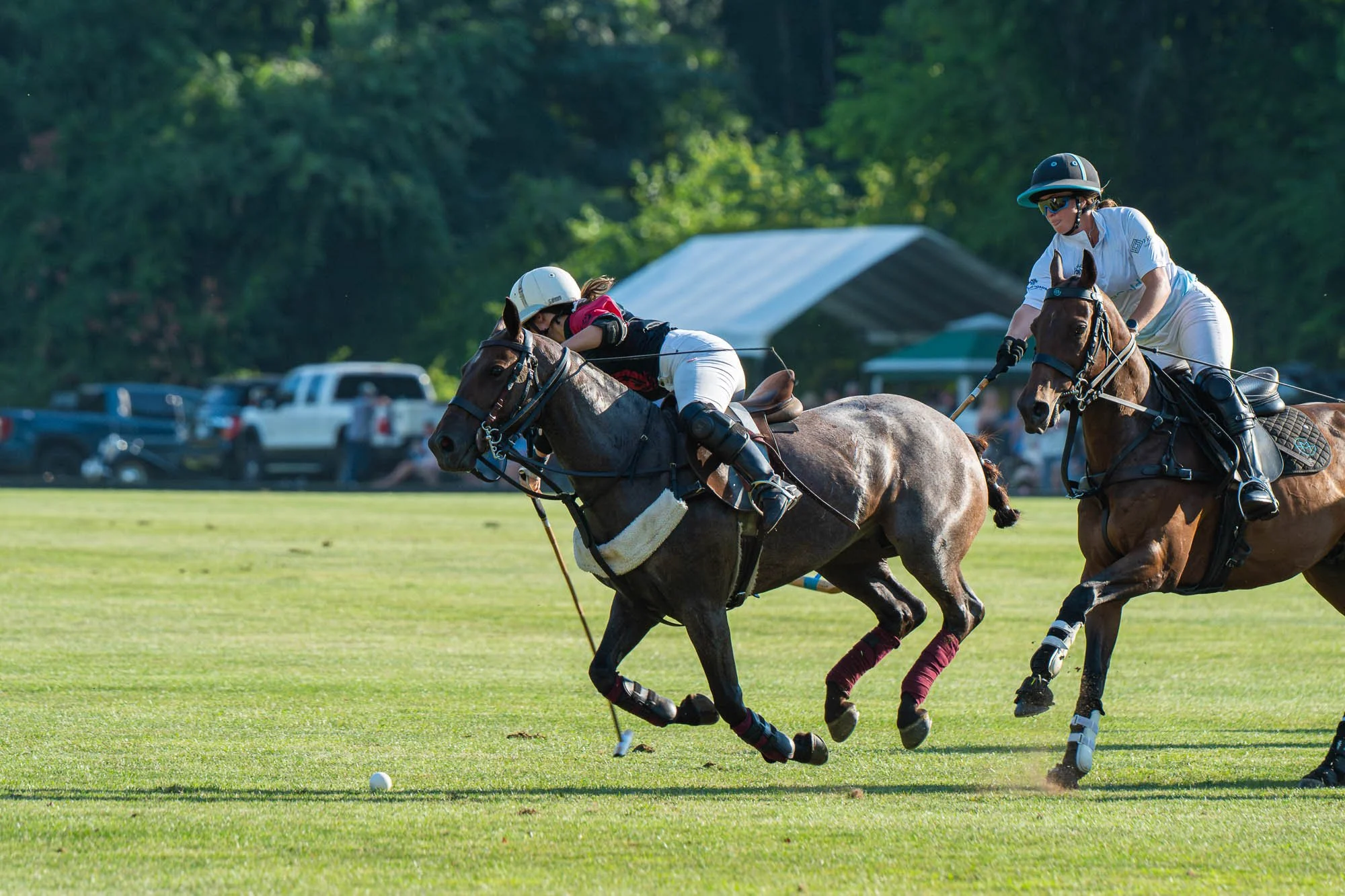 Two women playing polo on horseback with a green, outdoor field and parked cars in the background.