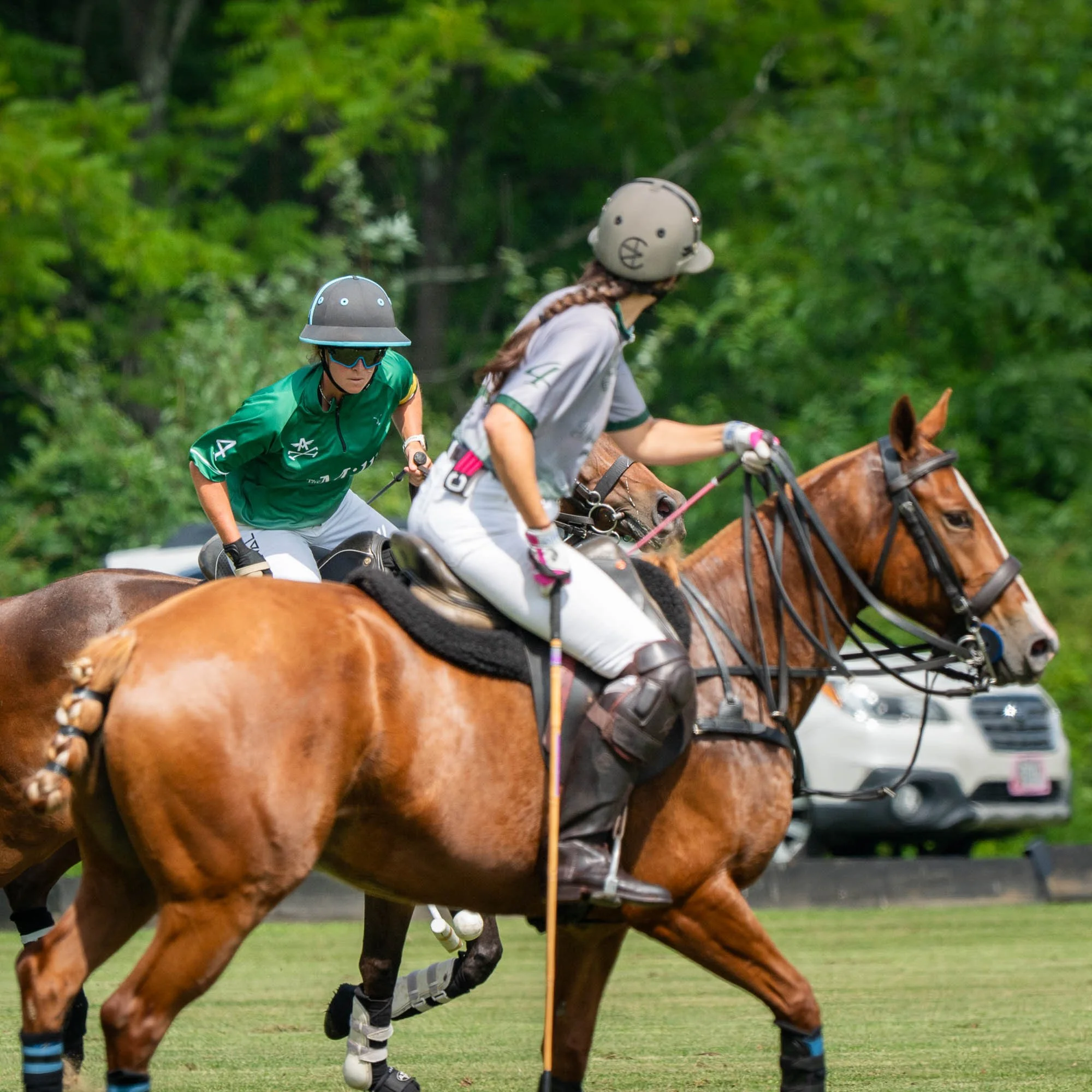 Two polo players riding horses compete in a match with green trees in the background. The player in the foreground wears a gray helmet and white attire, while the player behind wears a black helmet and green shirt.