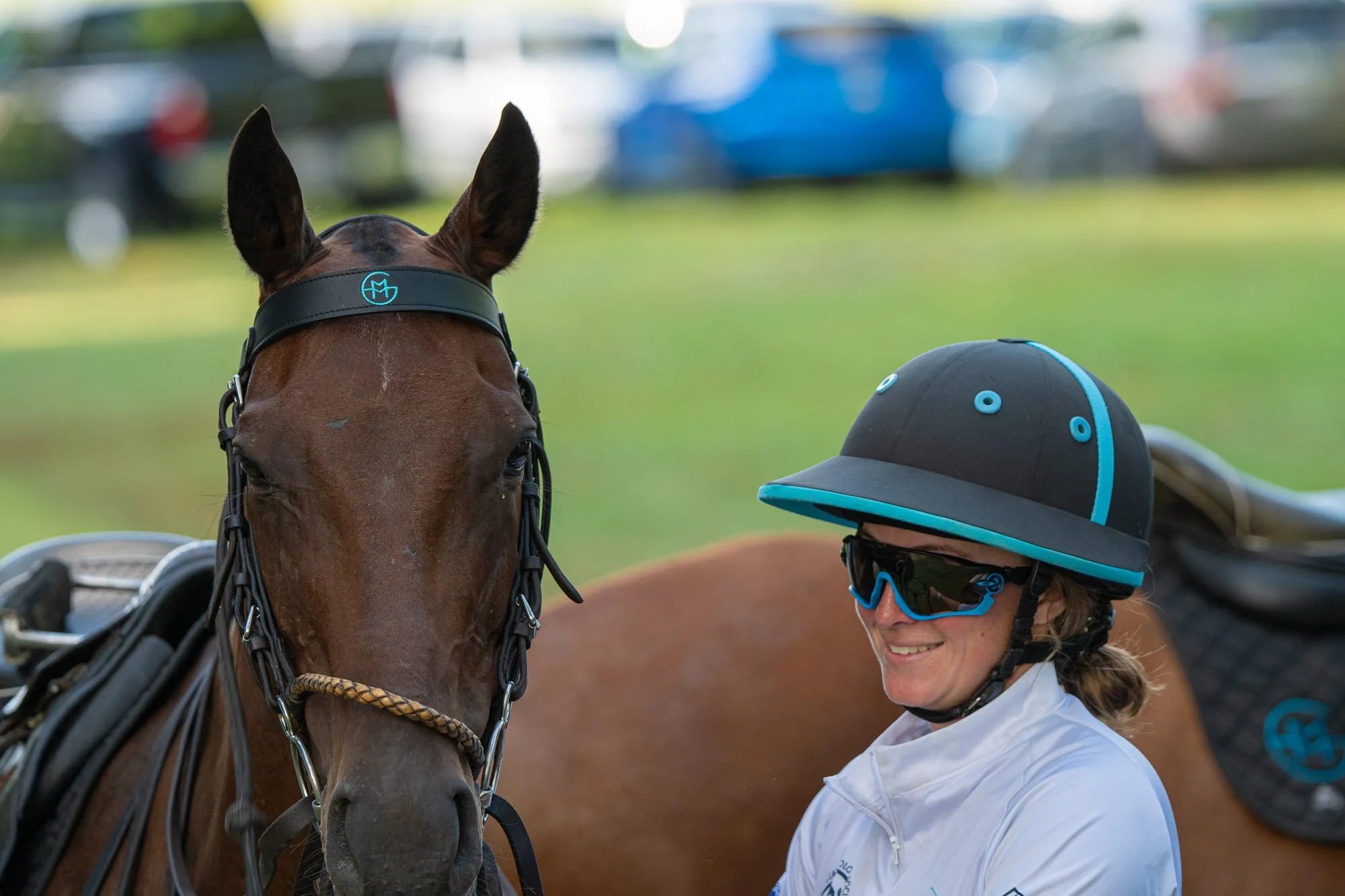 A woman in equestrian gear smiling, wearing a black and blue riding helmet with a blue logo, next to a brown horse with a black bridle and outdoor urban background with cars and grass.