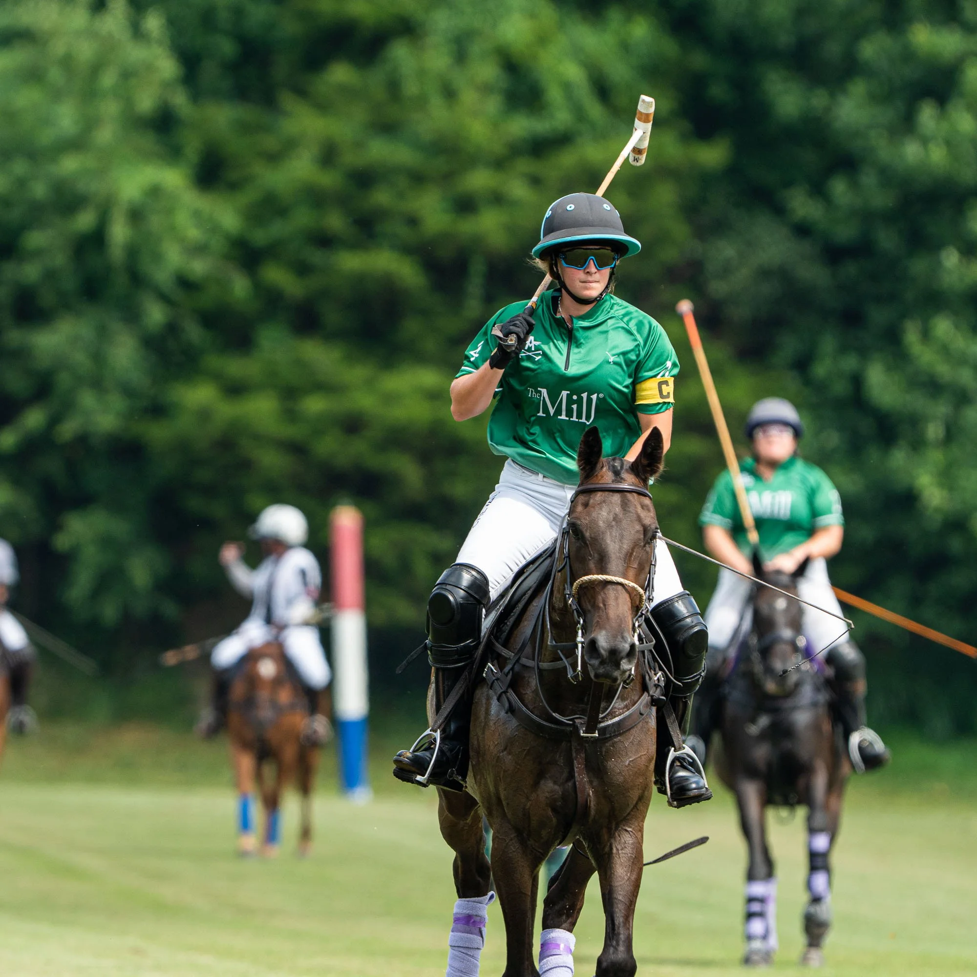 A person riding a horse during a polo match, wearing a green jersey with text that reads 'The Mill', black helmet, sunglasses, and gloves, with other players visible in the background.