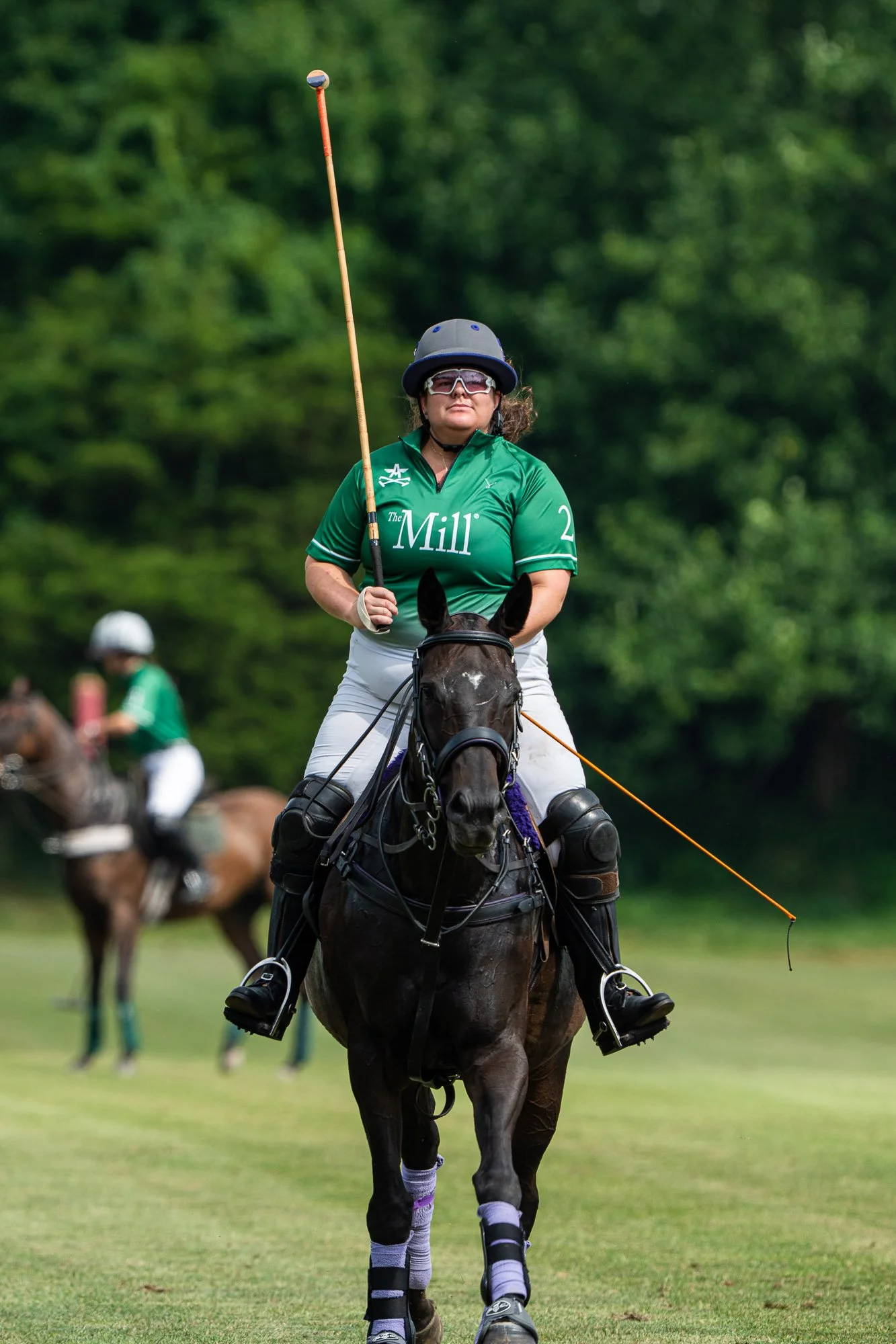 A woman riding a polo horse on a green field with trees in the background. She is wearing a helmet, sports glasses, a green shirt, and white riding pants. She holds a polo mallet in her right hand and a rein in her left.