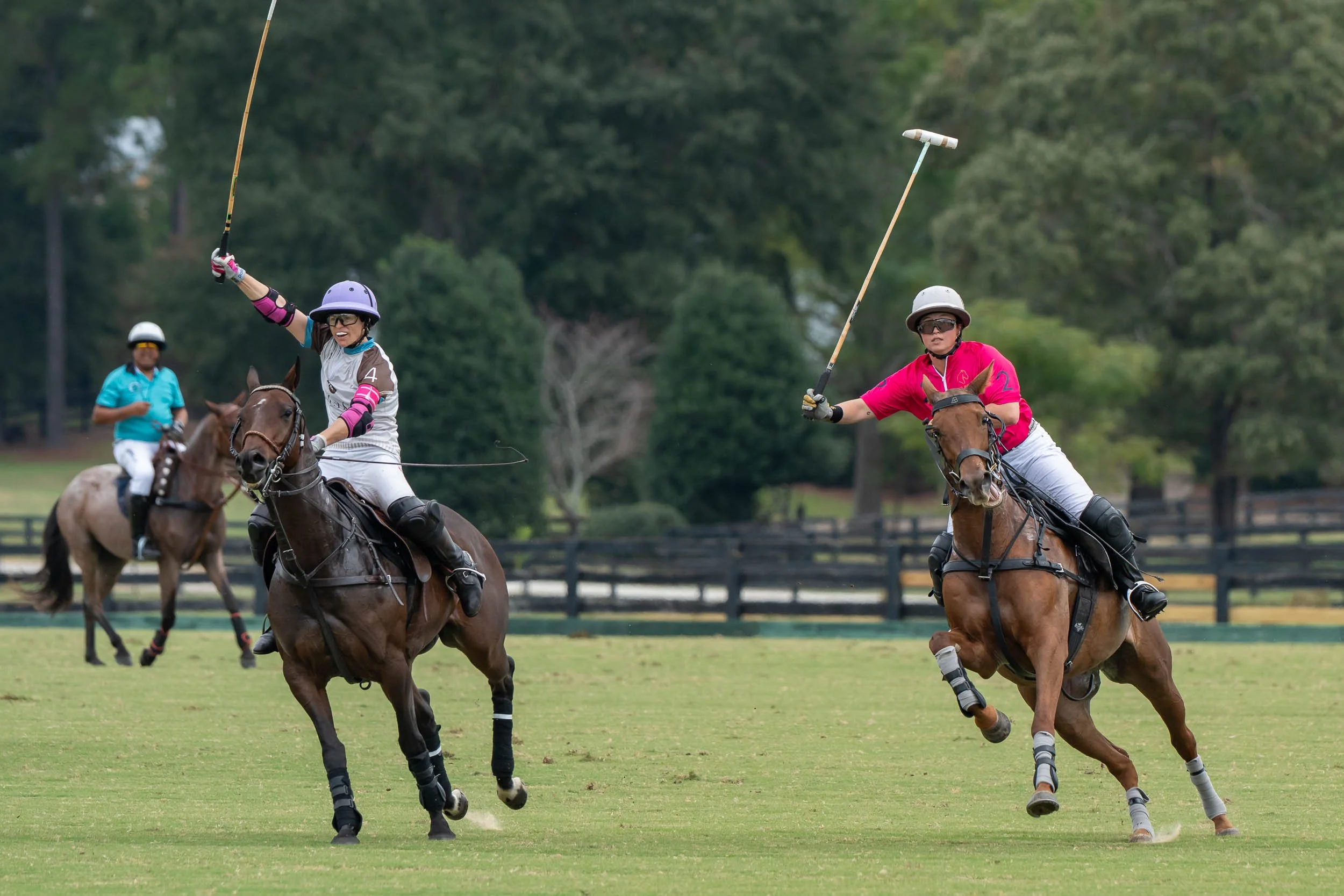 Polo players in helmets ride horses and swing mallets during a match on a grassy field.