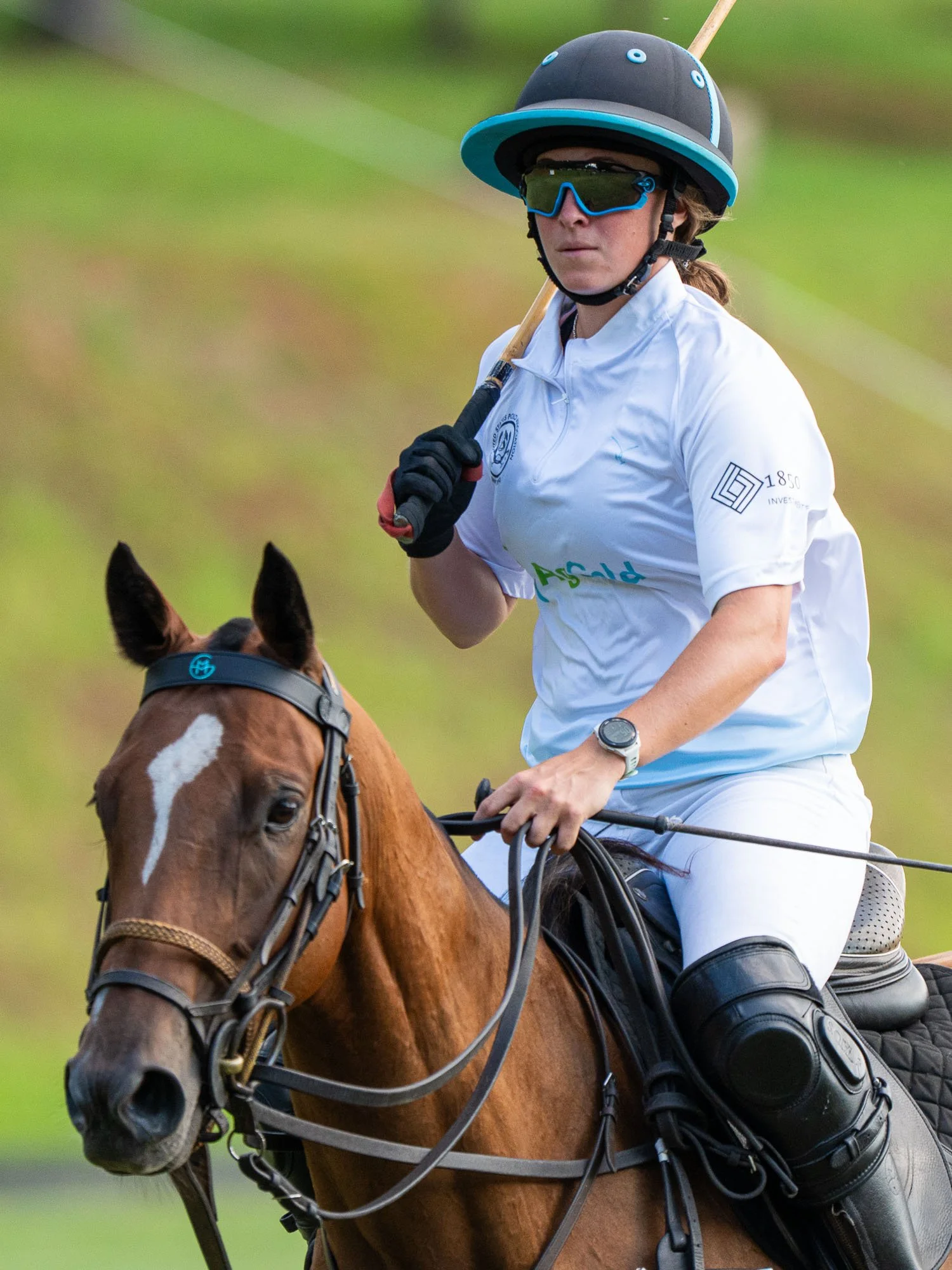 A woman wearing a helmet, sunglasses, and a white polo shirt riding a horse during a polo match.