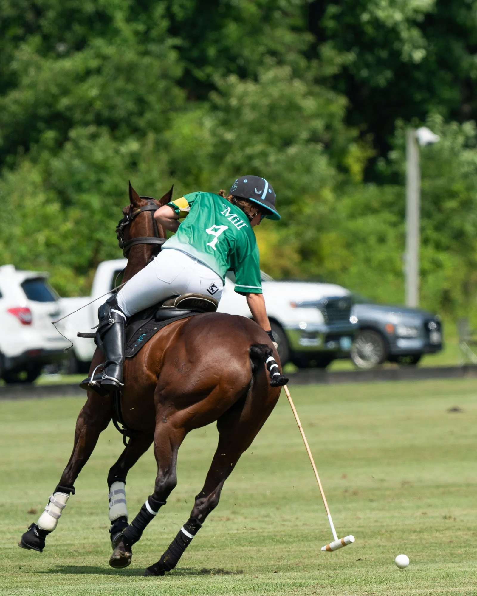 A polo player in a green jersey with the number 4, wearing a black helmet, rides a brown horse and skillfully hits a white ball with a mallet during a polo match on a grassy field with parked cars and green trees in the background.