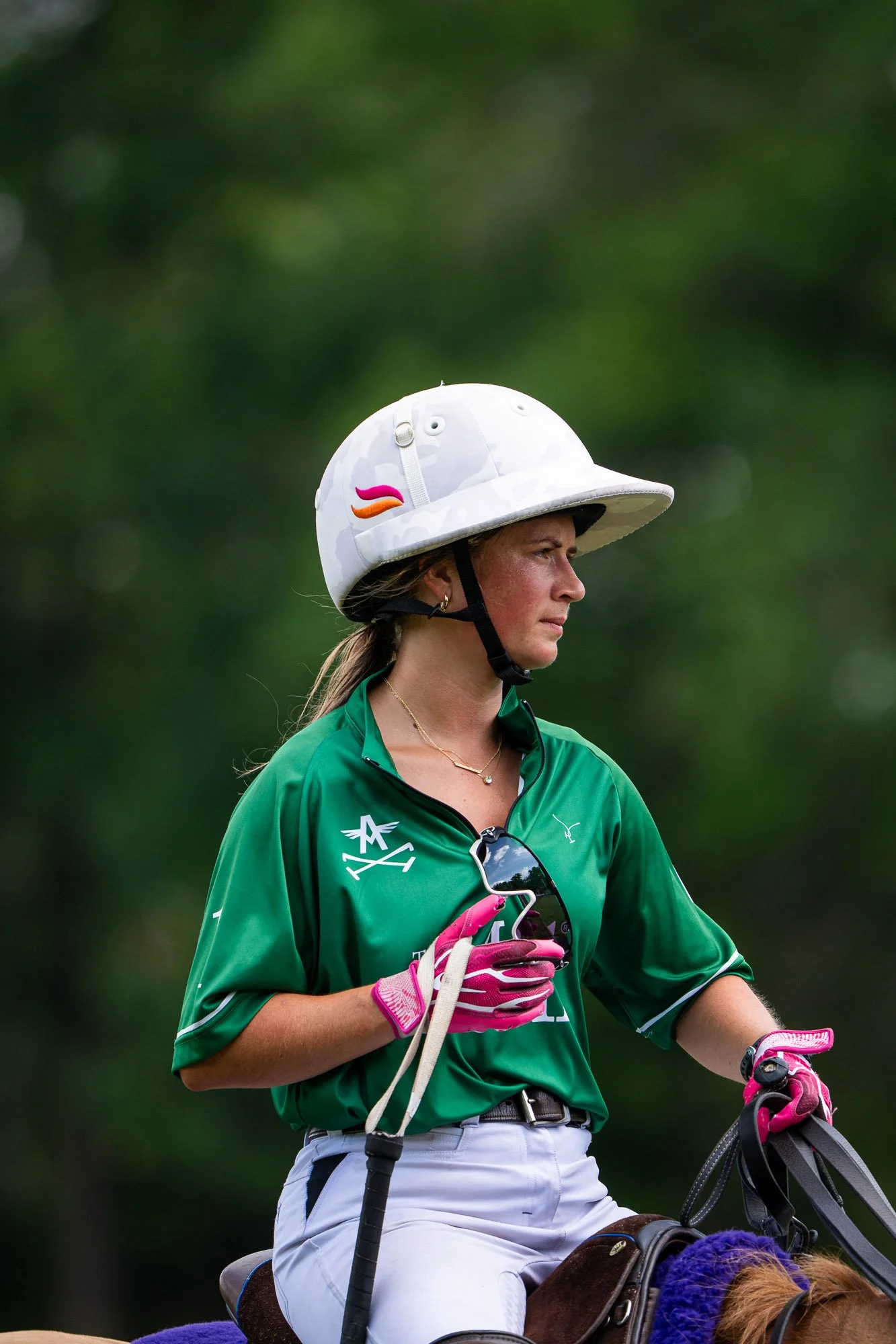 A woman wearing a white helmet with colorful accents and a green shirt is riding a horse. She is holding pink and gray gloves and a riding crop, with sunglasses hanging from her shirt. The background is blurred with green foliage.