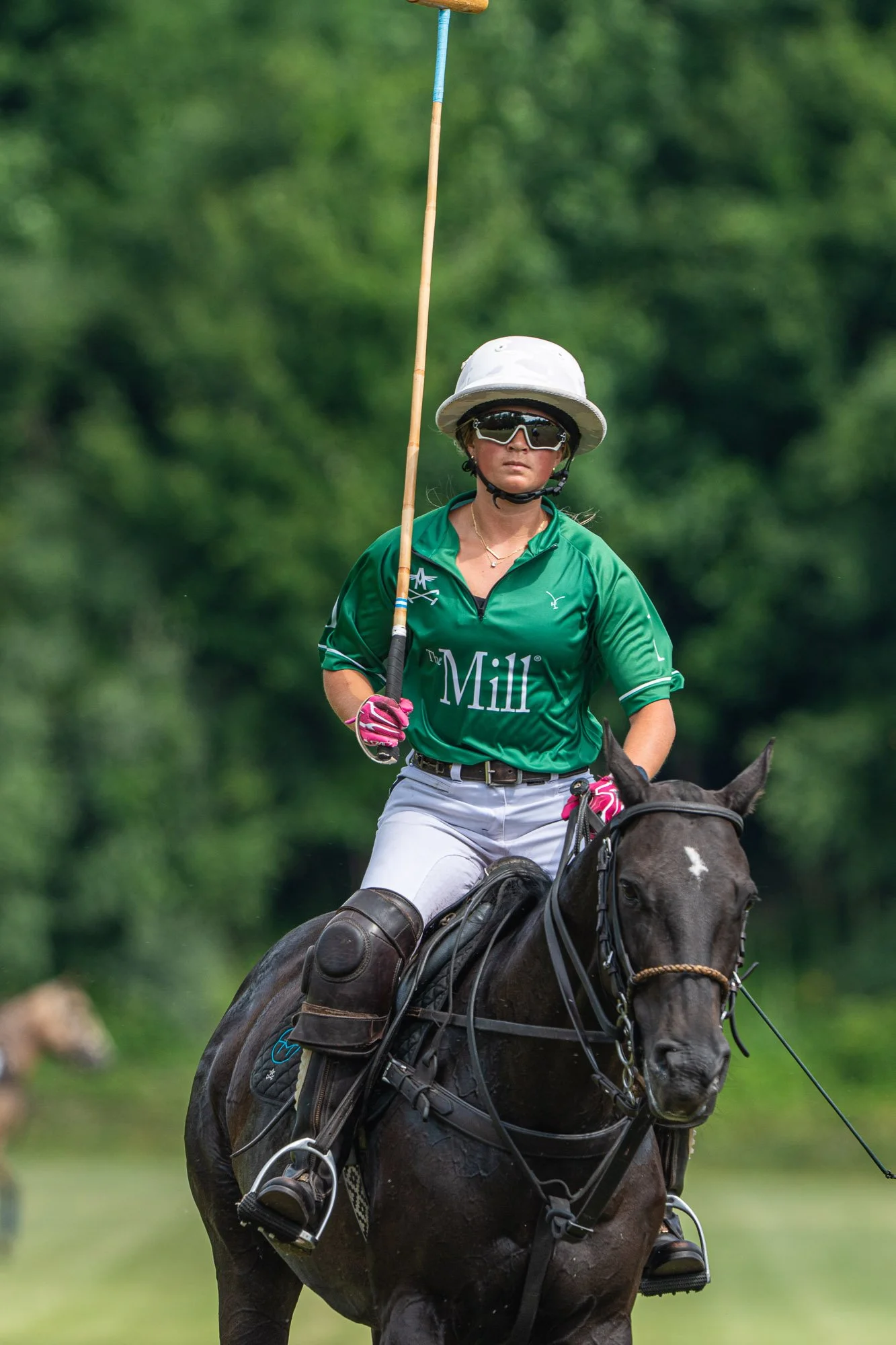 Female polo player riding a dark horse wearing a white hat, sunglasses, a green shirt with the word 'The Mill,' white pants, and pink gloves, holding a polo mallet. The background is blurred green trees.