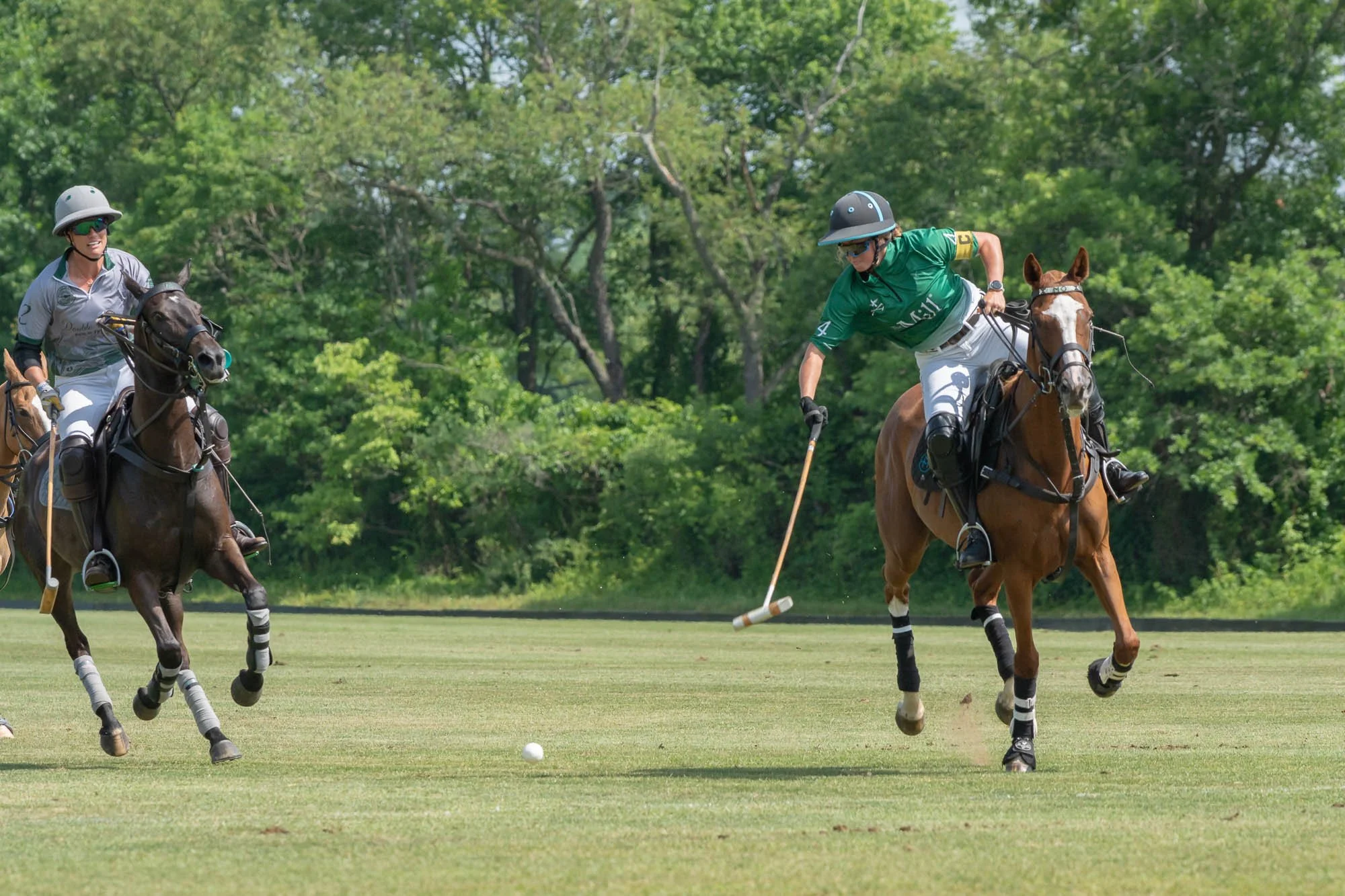 Two polo players riding horses are competing for the ball on a grassy field surrounded by trees. Both players are dressed in athletic gear and wearing helmets.