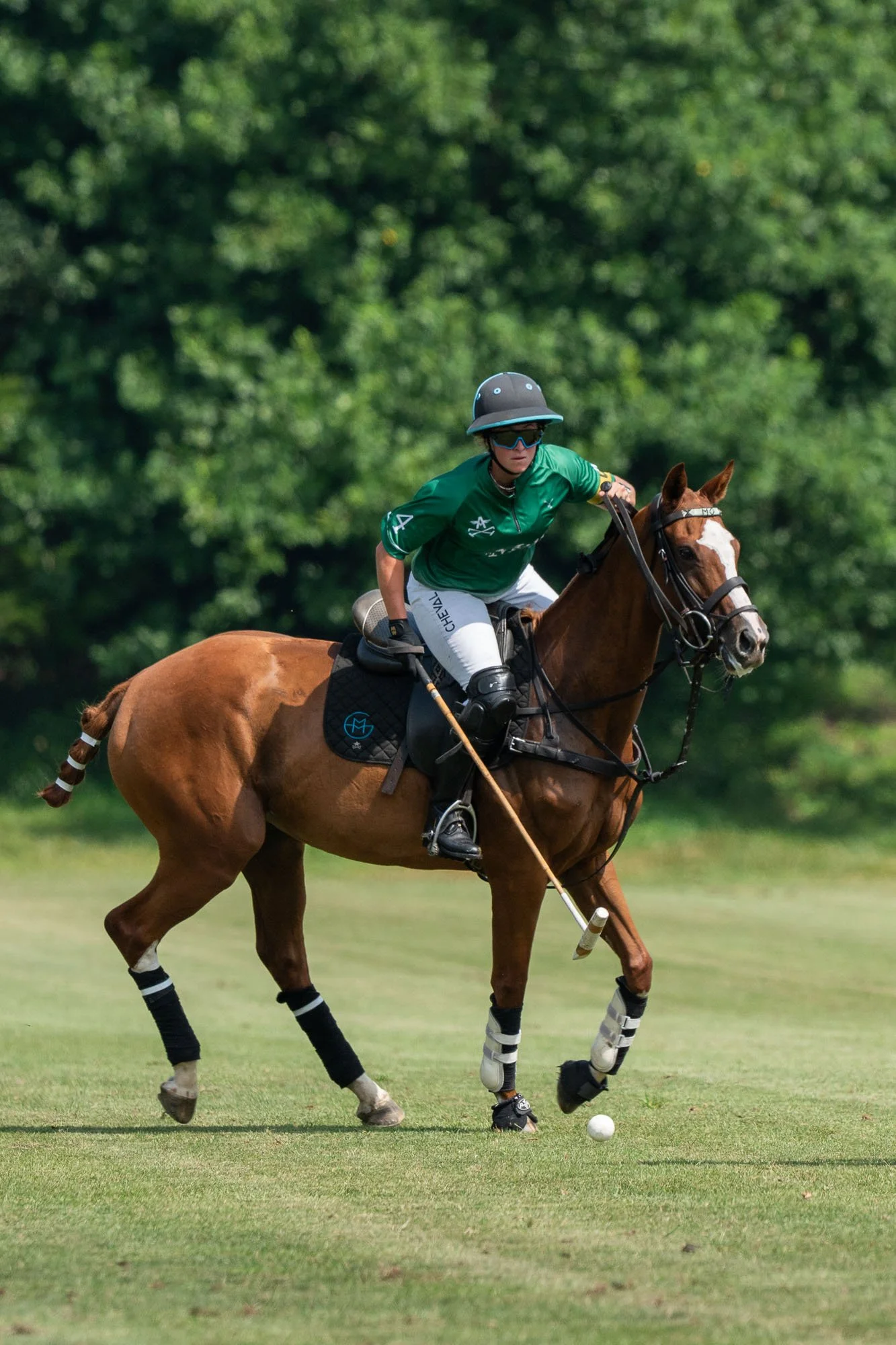 A Polo player riding a brown horse, wearing a green jersey, black helmet, sunglasses, and white pants, is hitting a white polo ball on a grassy field with a background of green trees.