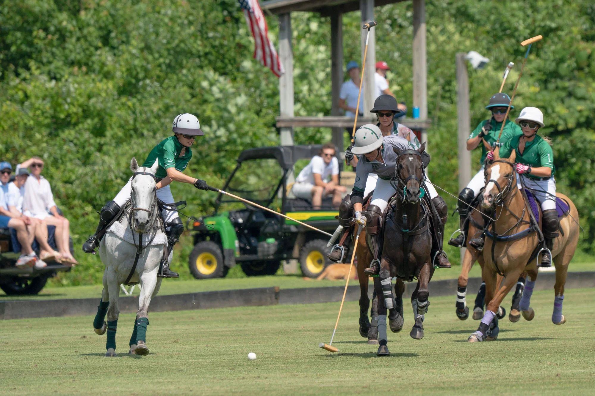 A group of polo players riding horses and hitting a polo ball during a match, with spectators watching and an American flag in the background.