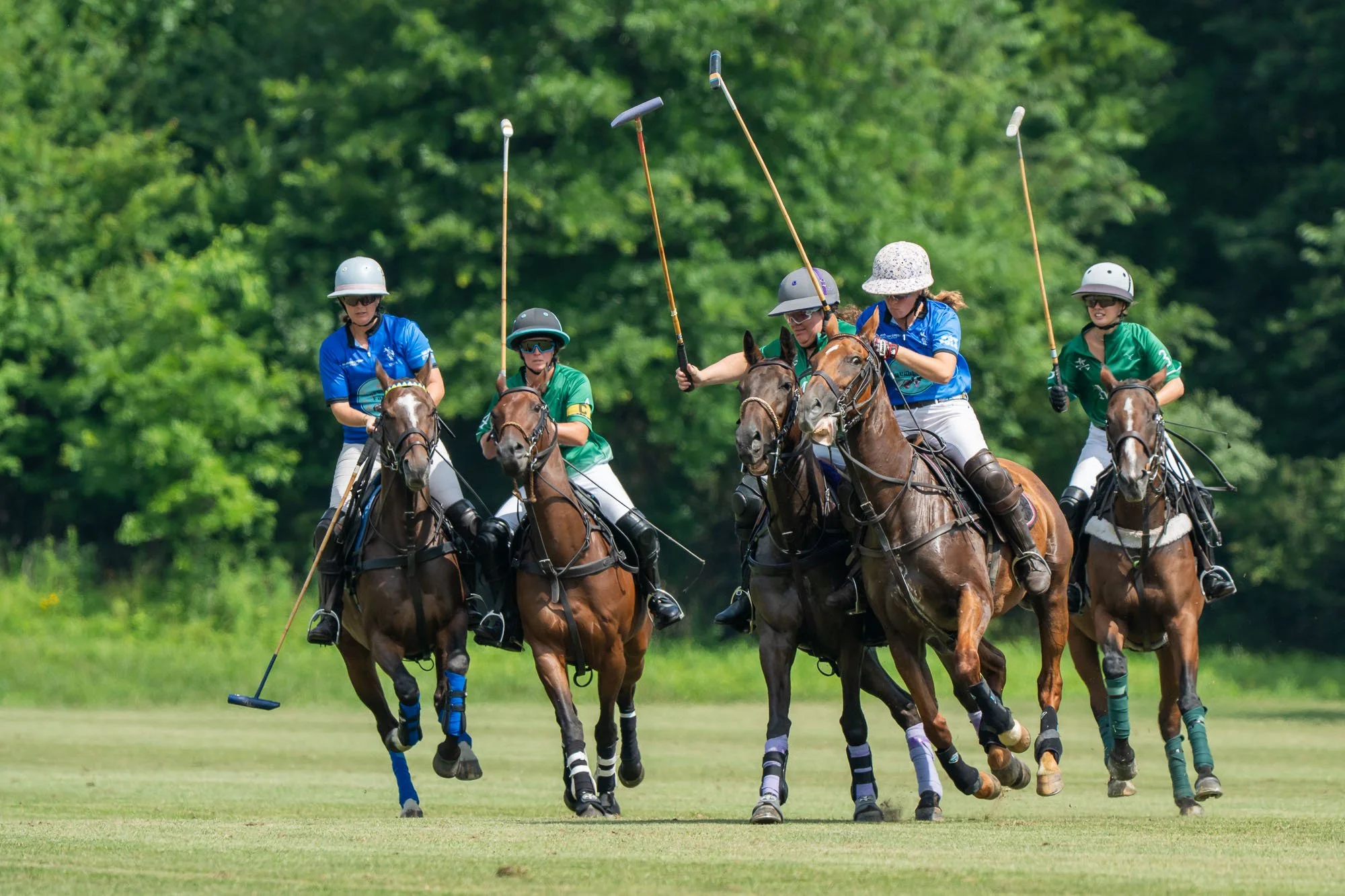 Six women paddle polo players riding horses on a lush green field during a game of polo, wearing helmets and colorful uniforms.