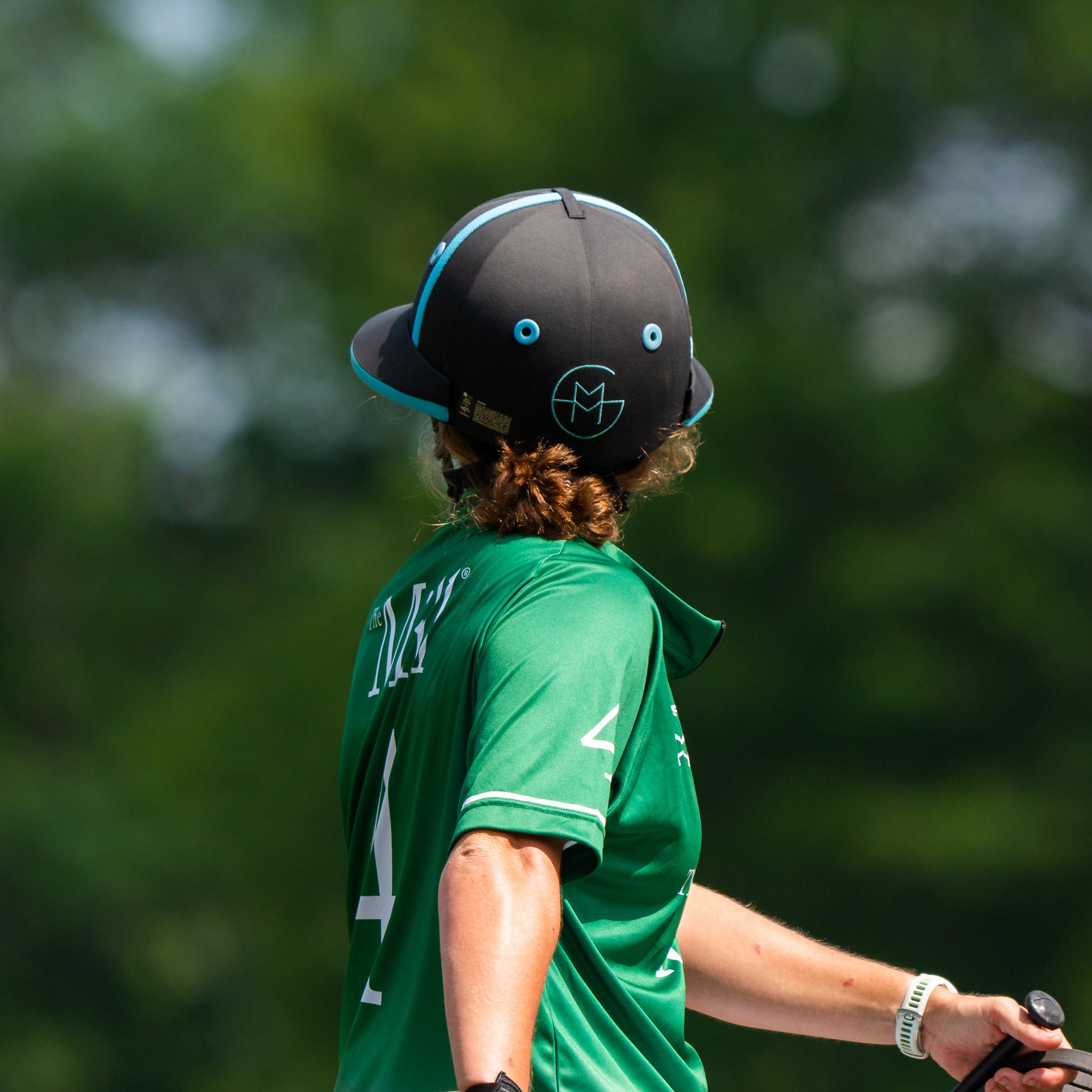 Close-up of a person wearing a black and blue helmet, green shirt, white wristwatch, and holding a golf club, standing outdoors with blurred green background.