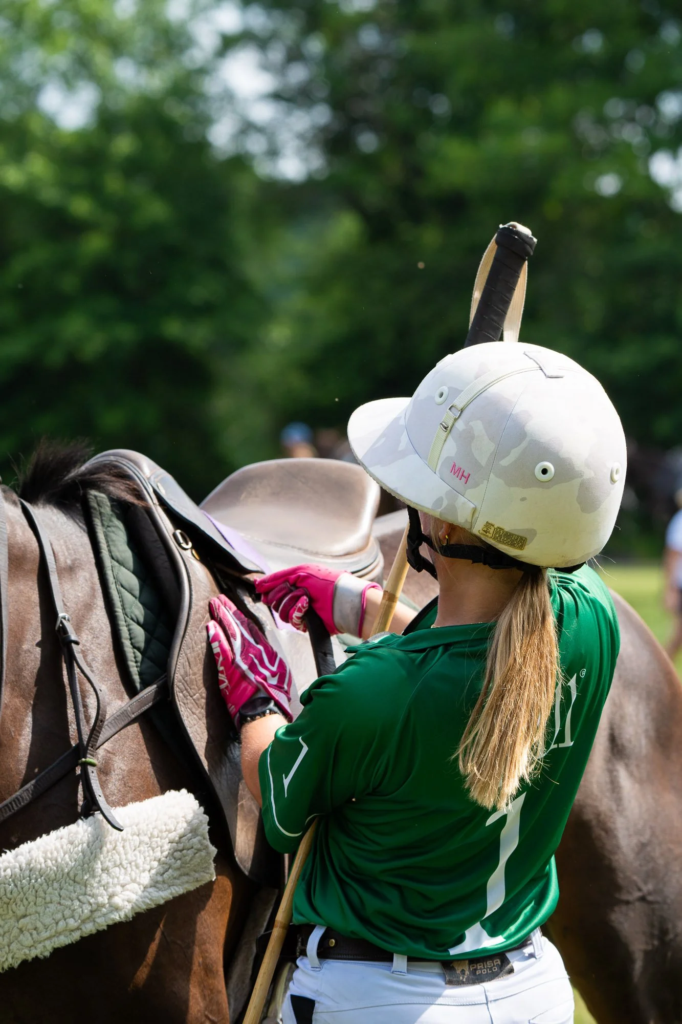 A woman wearing a helmet and green shirt grooming a horse with a brush outdoors.
