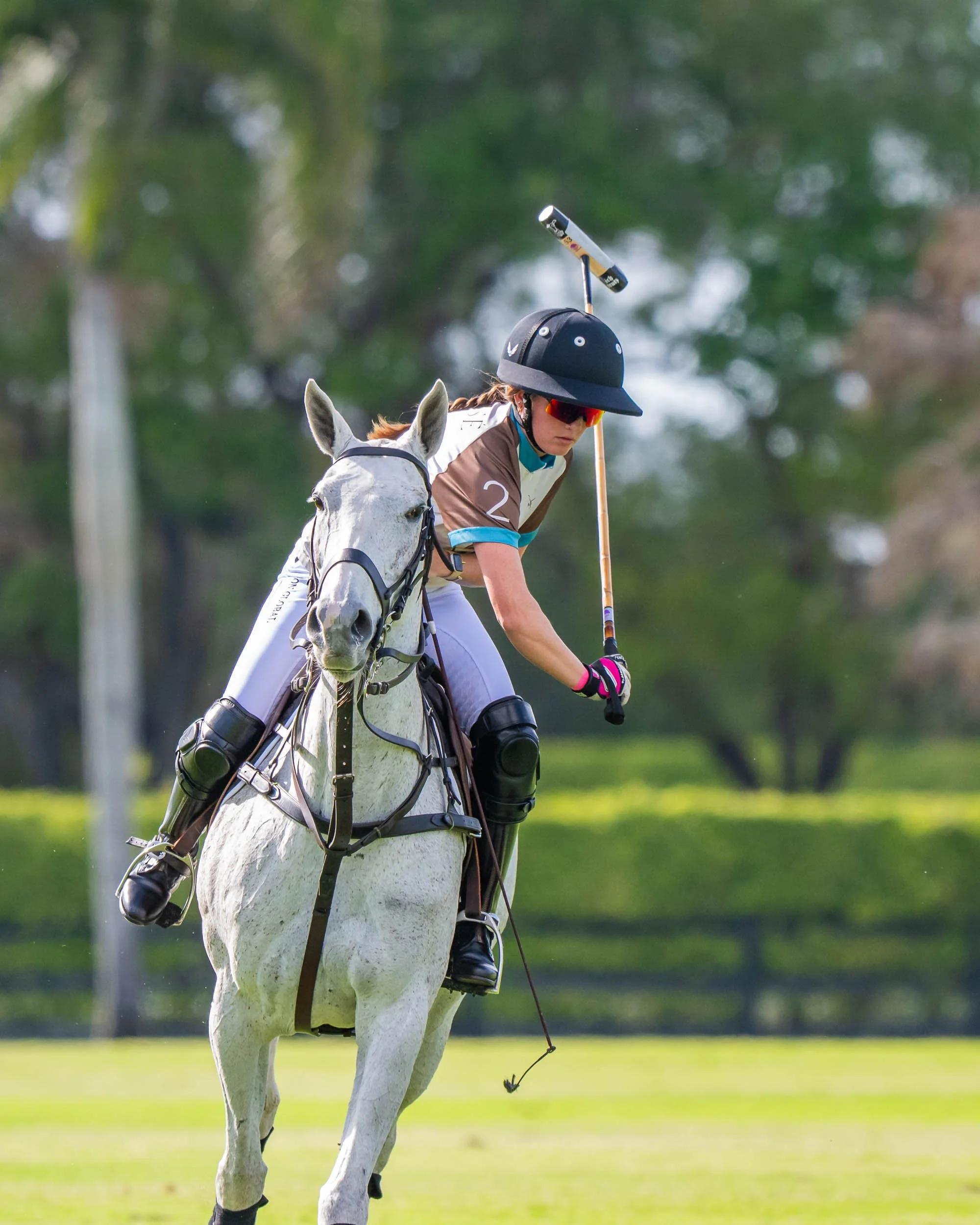 A person riding a white horse on a green field with trees in the background, wearing a black helmet, sunglasses, and casual riding attire.