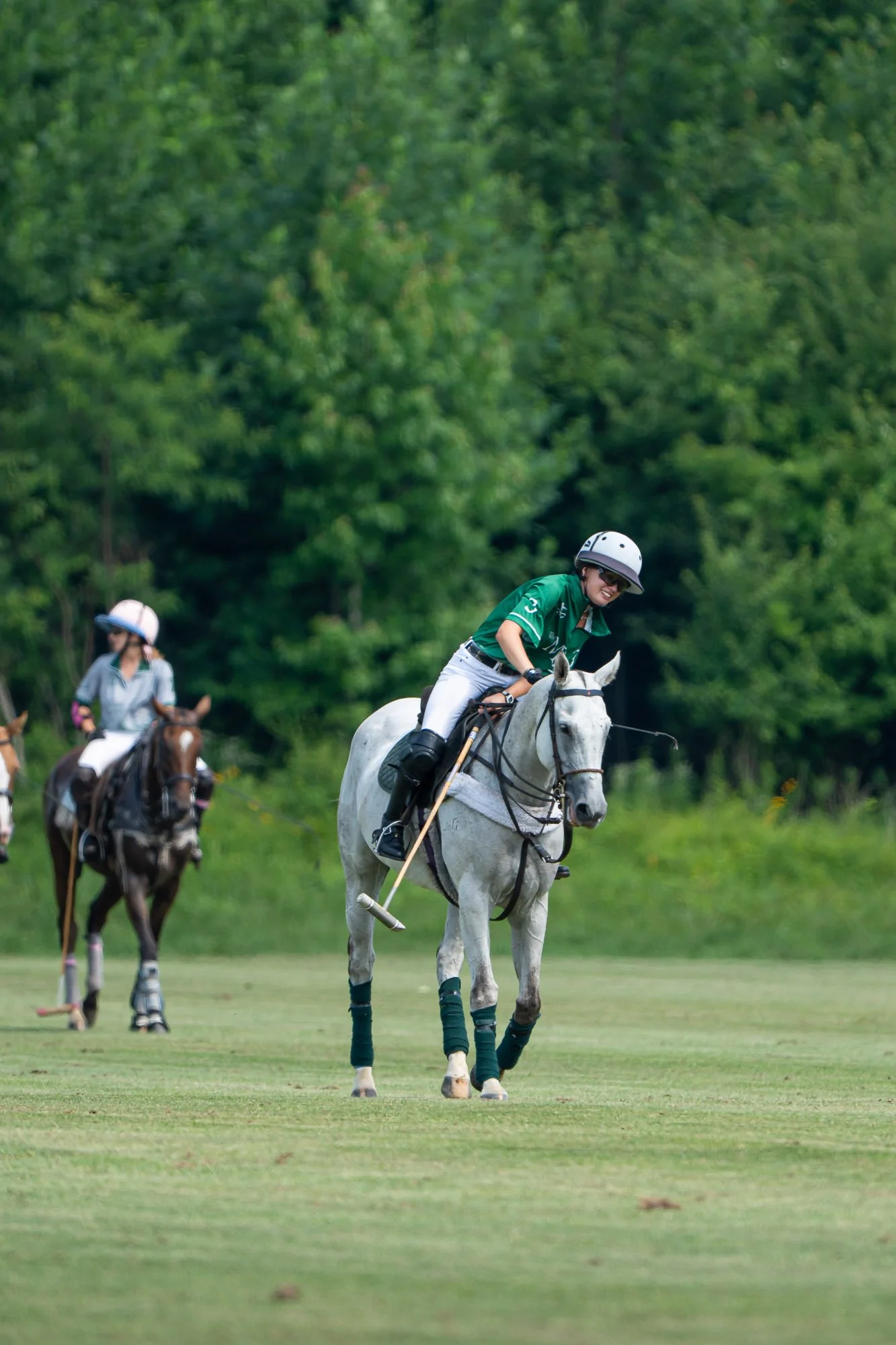 Two women riding horses in a green field with trees in the background. The woman in the foreground is smiling and wearing a green shirt, white pants, a helmet, and sunglasses.