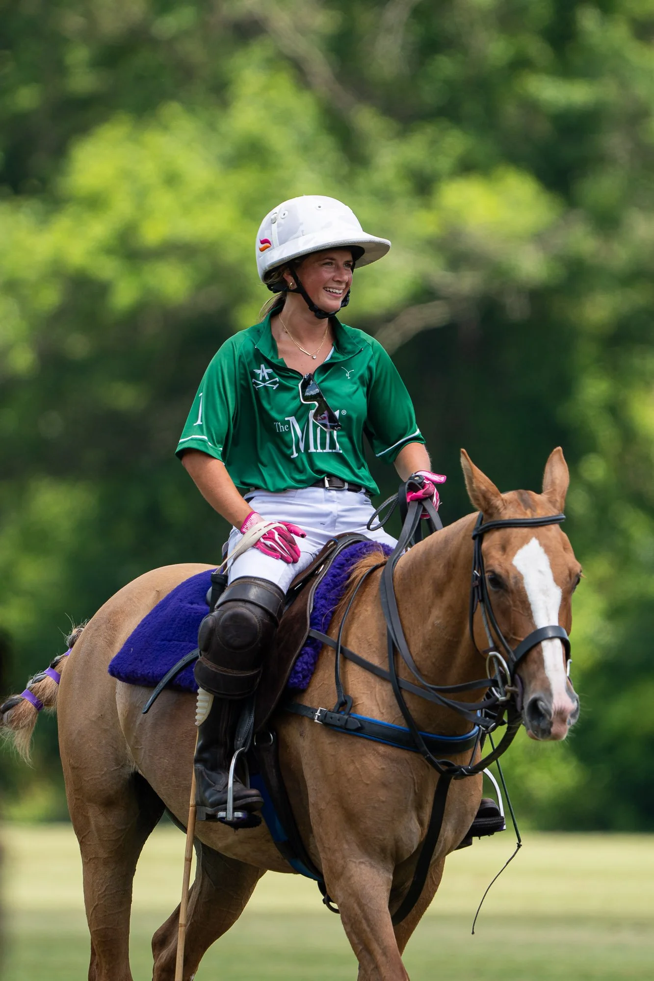 A woman riding a brown horse outdoors, wearing a green shirt, white helmet, pink gloves, and sunglasses, with a smiling expression.