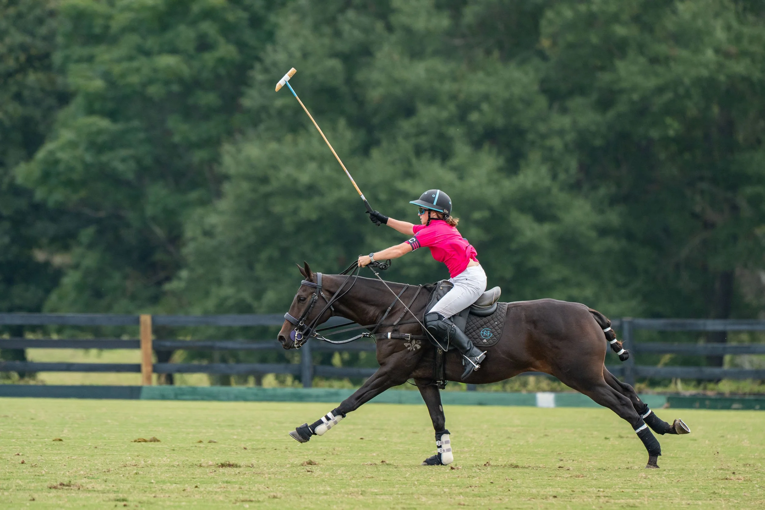 Polo player in pink shirt riding a galloping horse and swinging a mallet on a grassy polo field