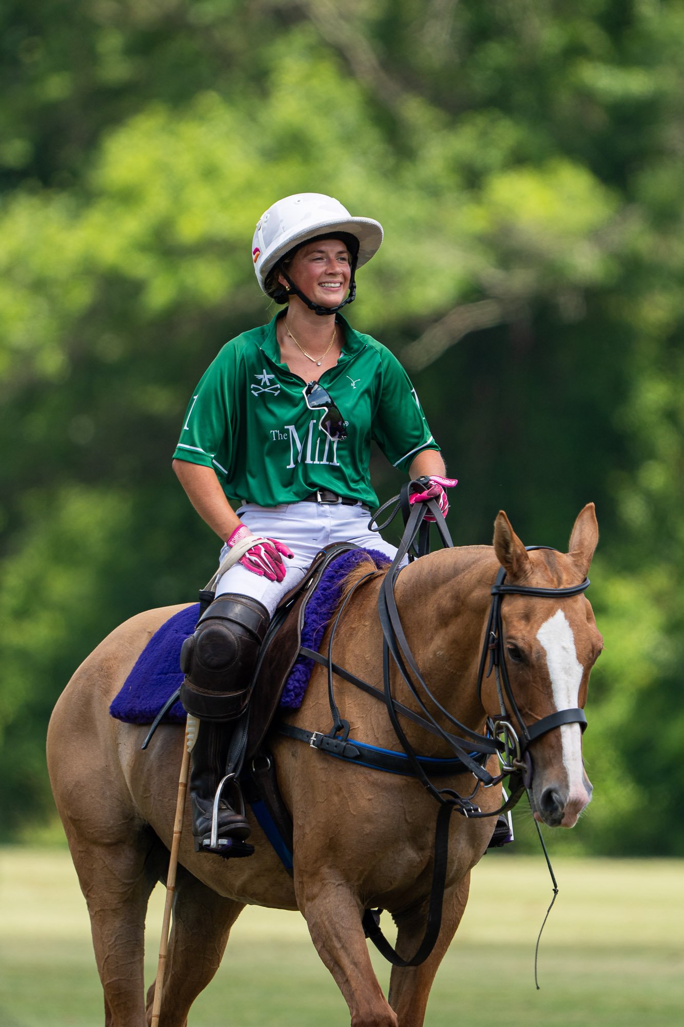 A woman wearing a green equestrian outfit and a white helmet riding a brown horse with a purple saddle pad in a lush green outdoor setting.