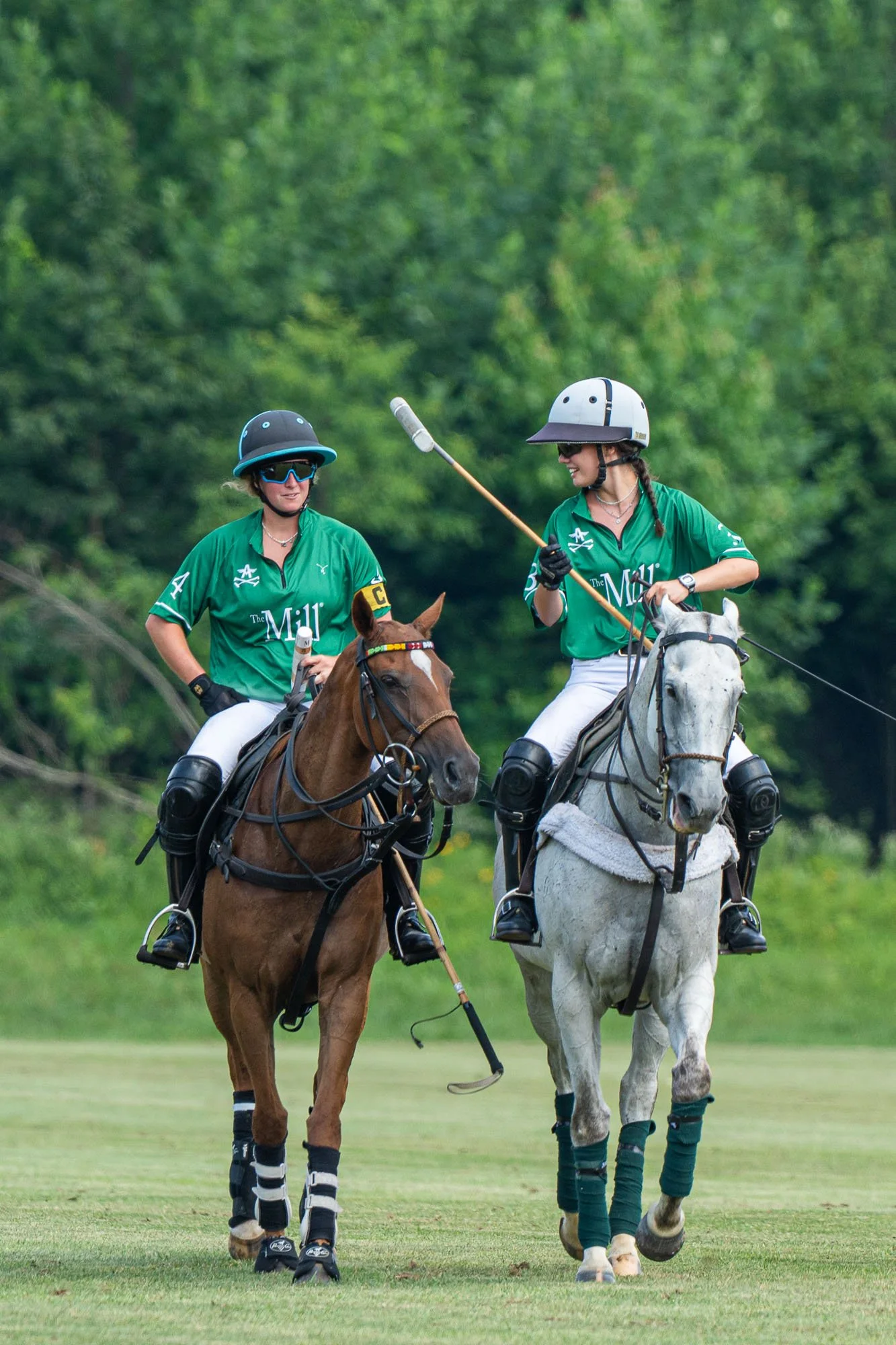 Two women in green polo shirts riding horses on a golf course, with a background of green trees. One woman is wearing bicycle gear including a helmet and sunglasses, and the other is wearing a helmet and glove, with a golf club in her hand.