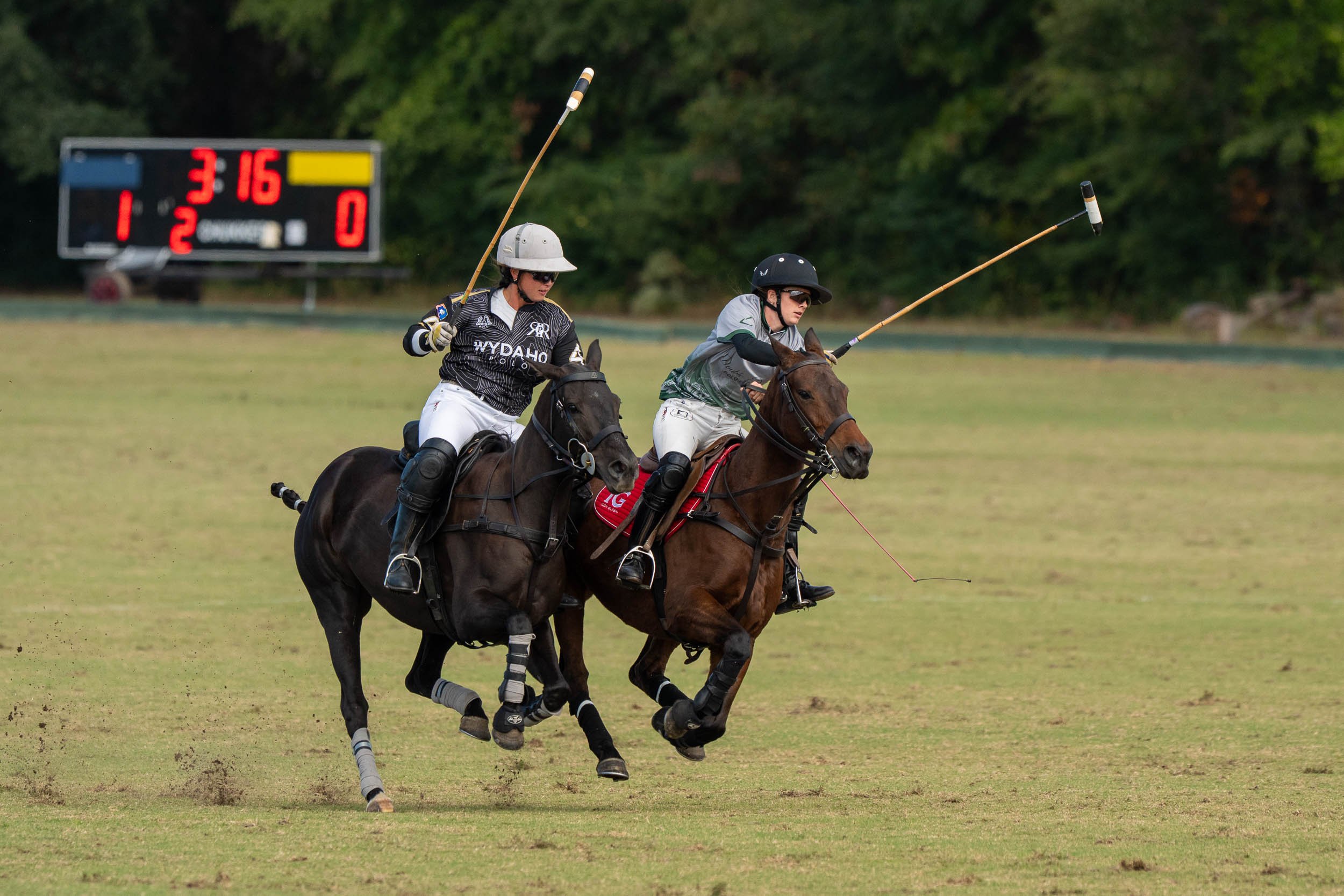 Two polo players on horseback competing on a grassy field, with mallets raised and a scoreboard in the background.
