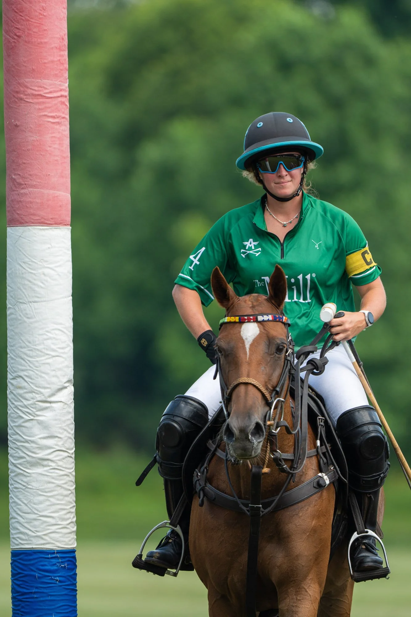 A woman wearing a green sports jersey, helmet, and sunglasses is riding a brown horse during a polo game. She has a yellow captain's armband and is holding a mallet, with a green blurred background.