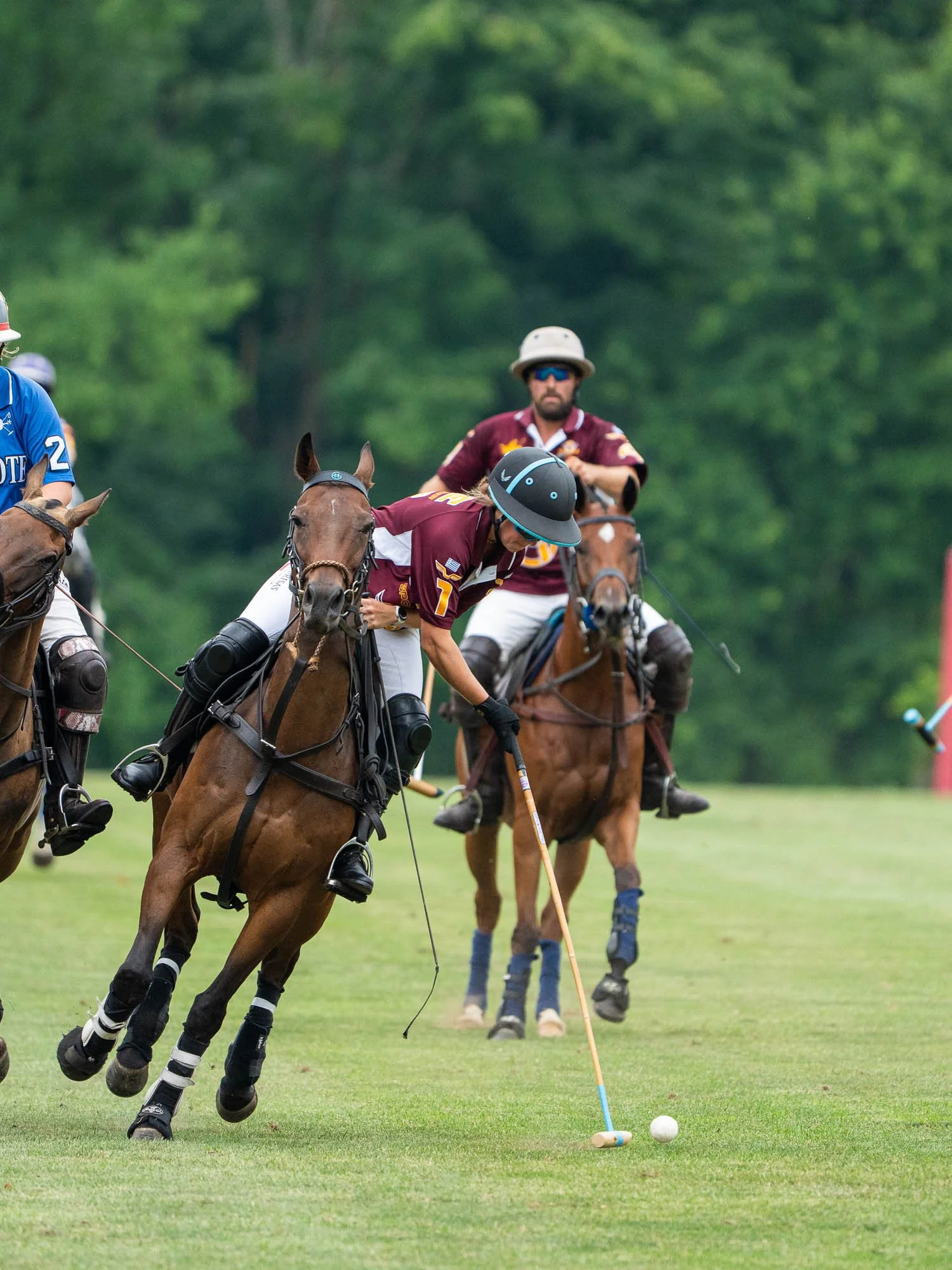 A polo match with players on horseback, one player bending down to hit the ball with a mallet on a green field, trees in the background.