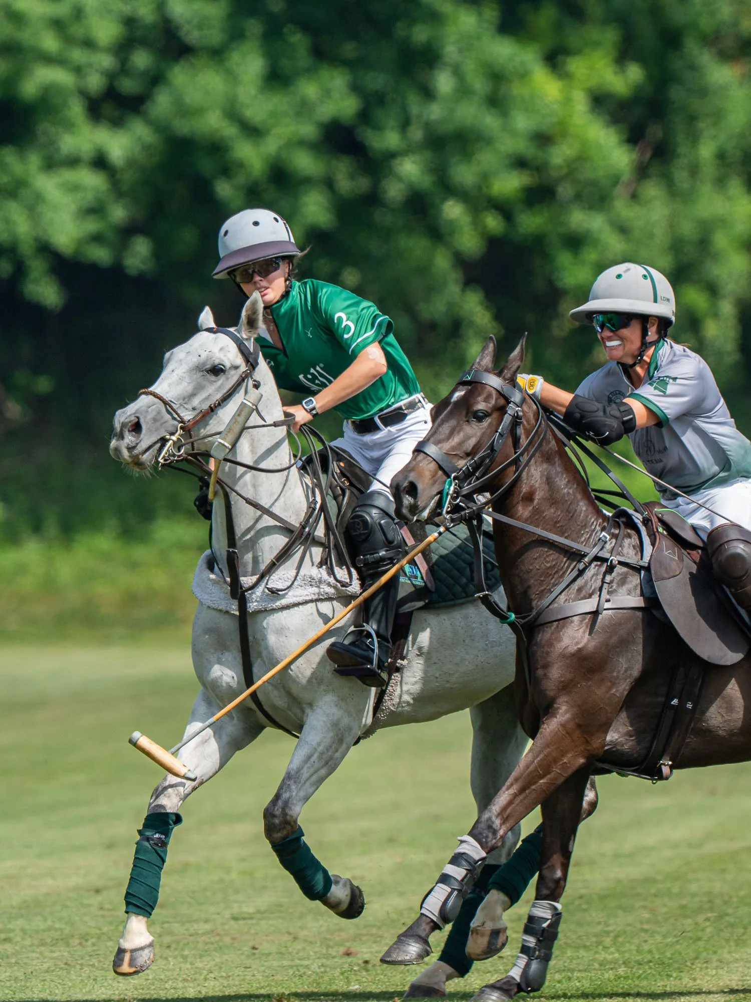 Two women playing polo on horses, both wearing helmets and sunglasses, with a green background.