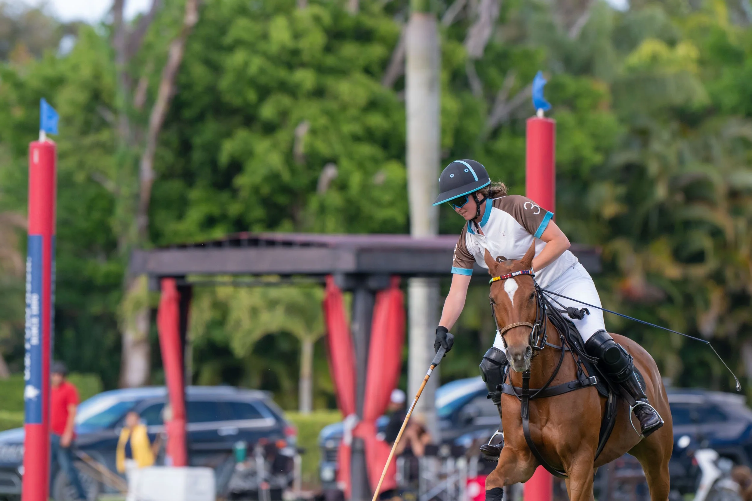 A woman riding a horse at a polo match, wearing a helmet and sunglasses, with trees and spectators in the background.
