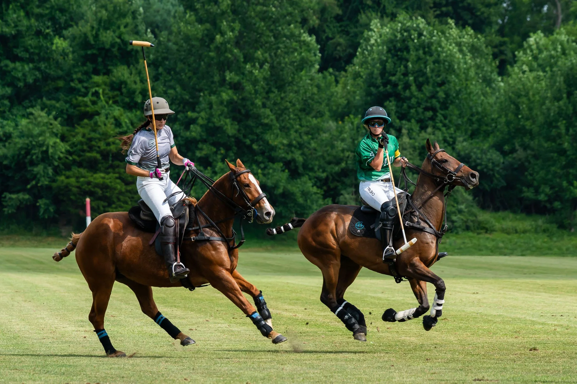 Two women riding horses on a grassy field, dressed in water polo uniforms, wearing helmets and sunglasses, with a background of green trees.