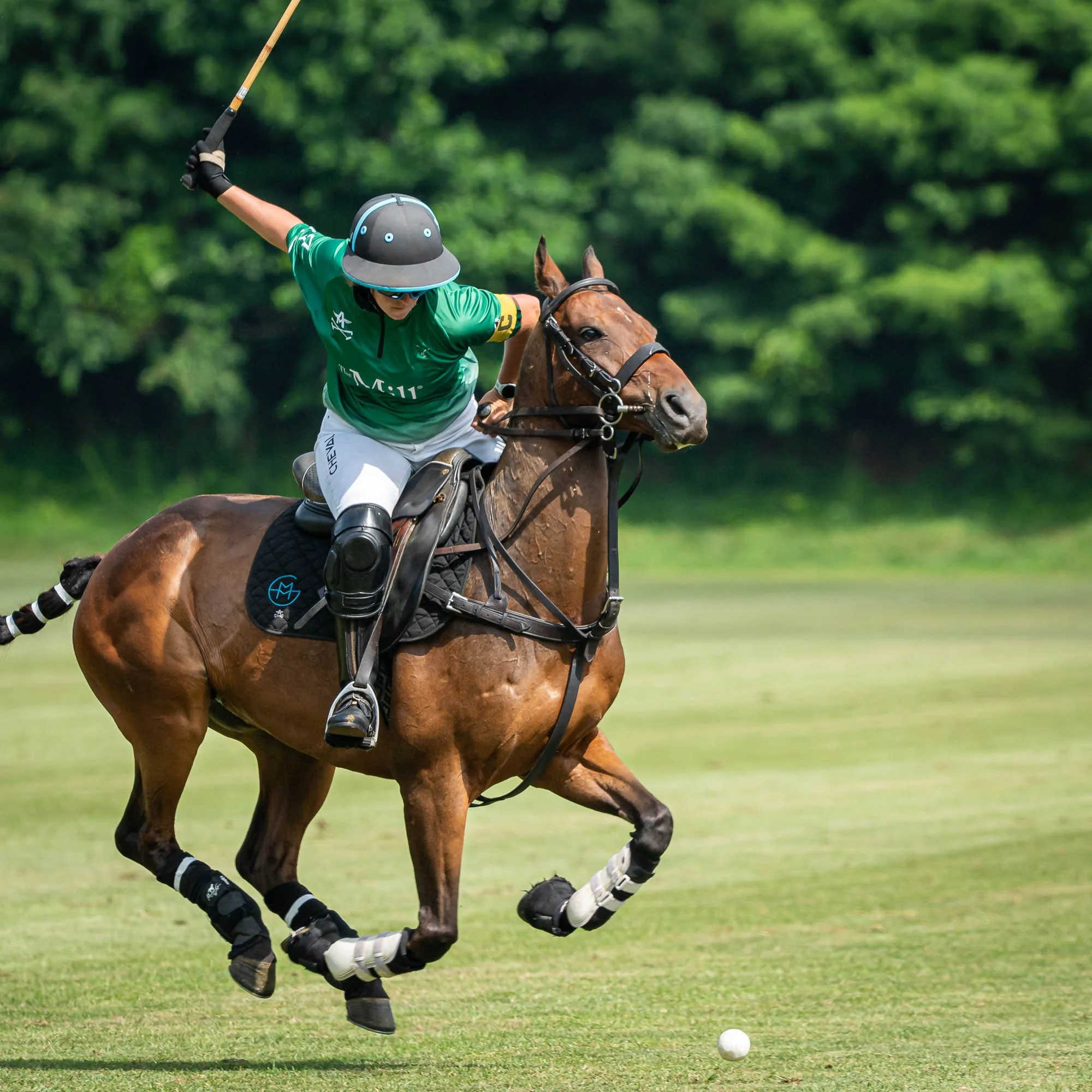 A polo player on a brown horse with a black saddle and gear, wearing a teal shirt, white pants, a black helmet, and sunglasses, reaching for a white polo ball on a grassy field, with trees in the background.