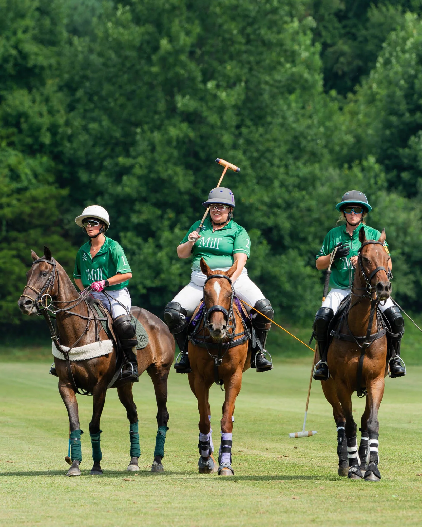 Three women playing polo on horseback on a lush green field with trees in the background. They are wearing green shirts, helmets, and sunglasses, and are holding polo mallets.