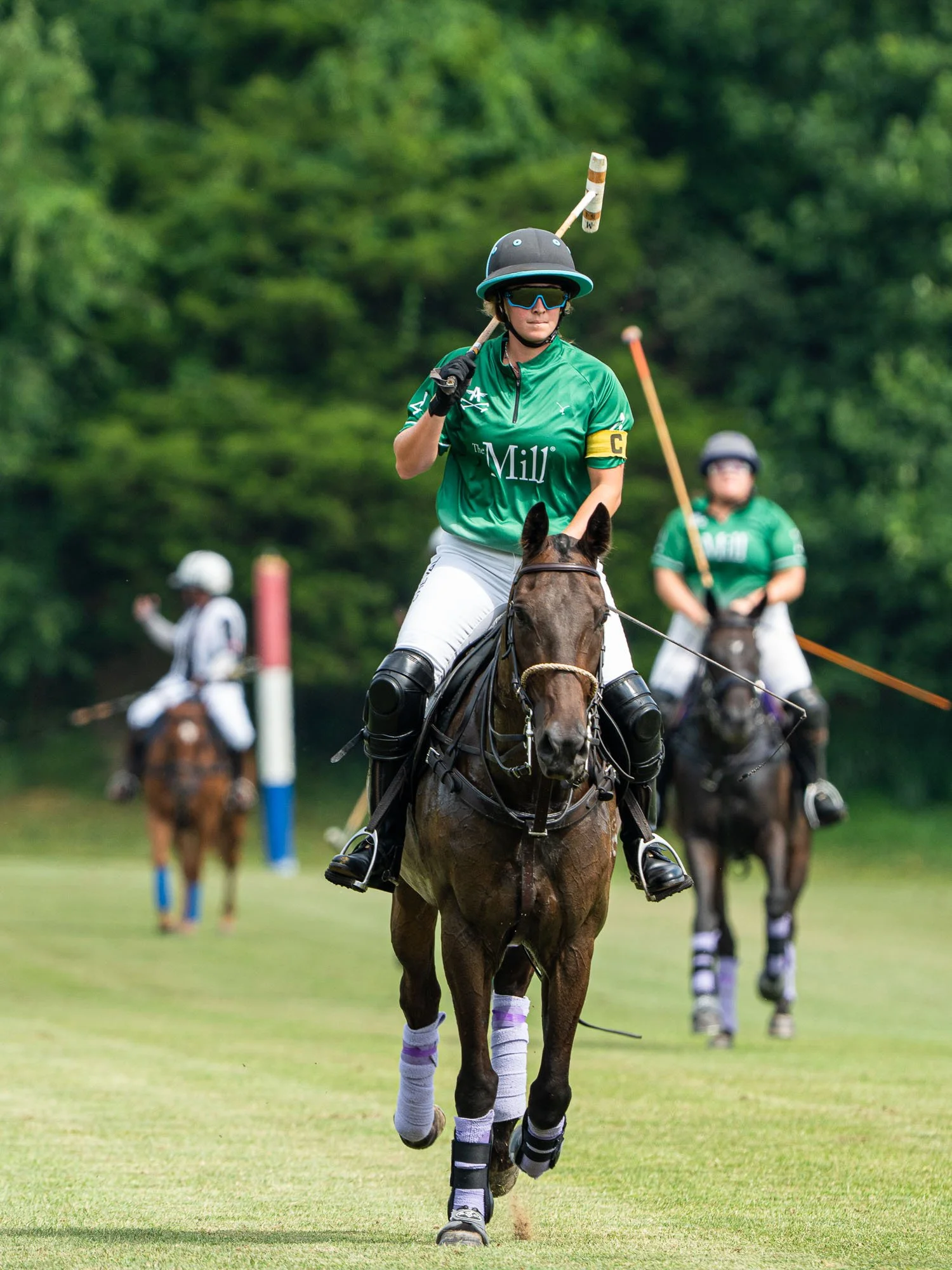 A polo player wearing a green jersey, riding a horse on a polo field, with other players in the background.