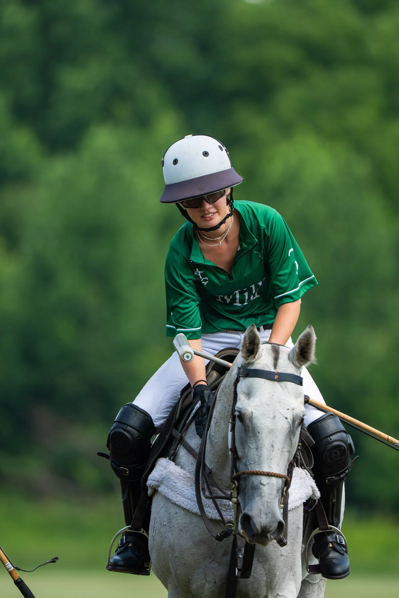 Person riding a white horse, wearing a green sports jersey, helmet, sunglasses, and gloves, with a lush green background.