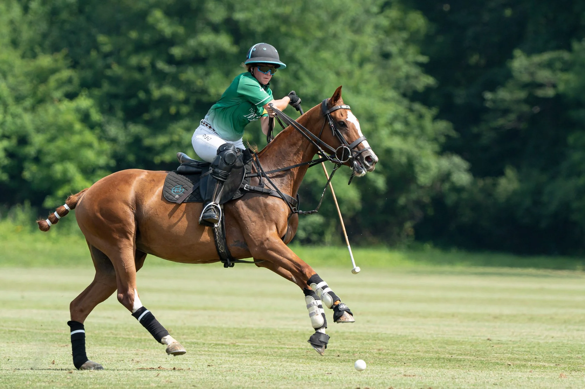 A polo player riding a brown horse on a grassy field, wearing a green jersey, helmet, and sunglasses, with a polo mallet in hand, focused on hitting a ball.