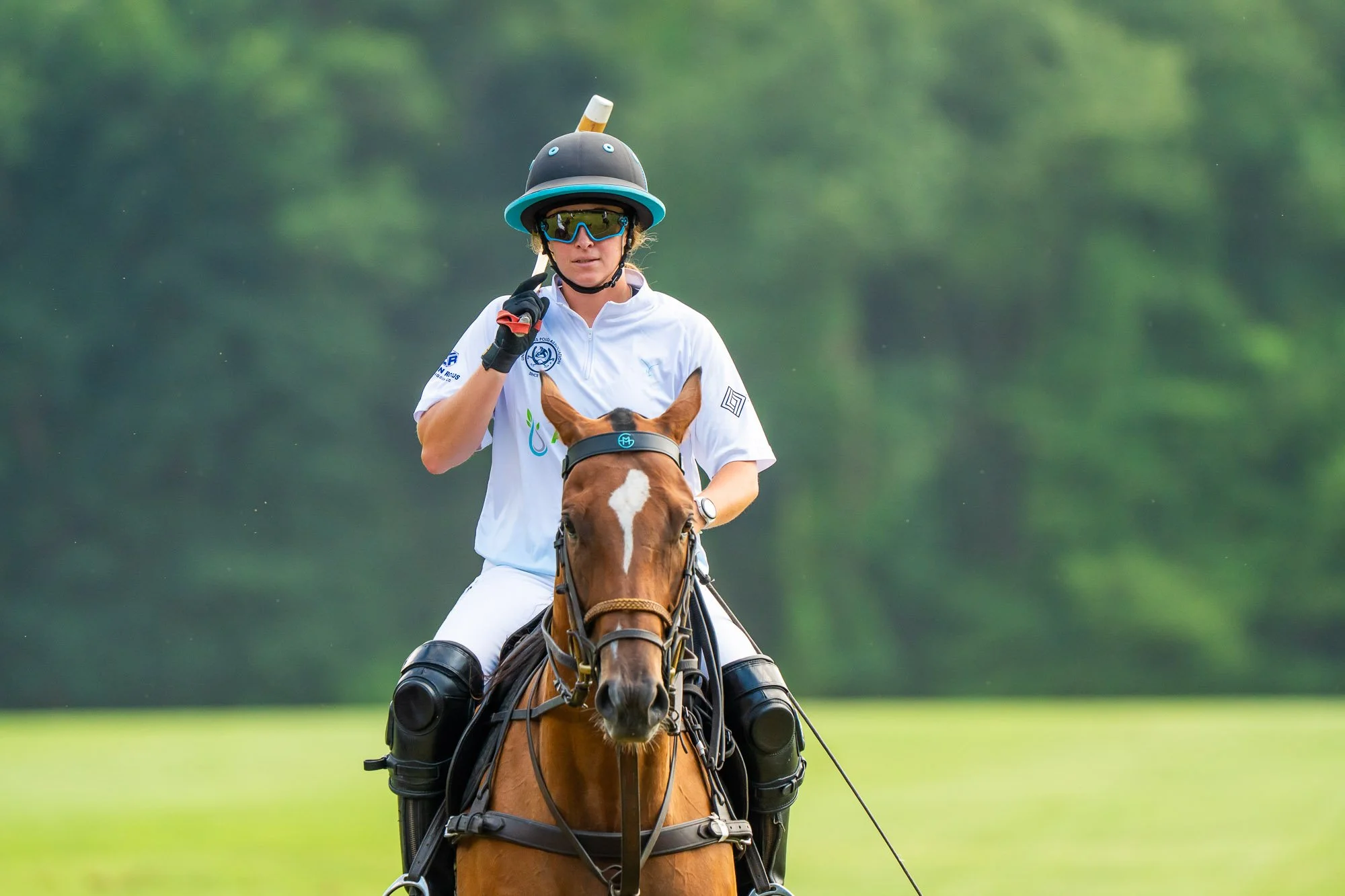 Woman riding a horse outdoors, wearing white sports gear, protective glasses, and a helmet with a bottle attached on top, with trees in the background.