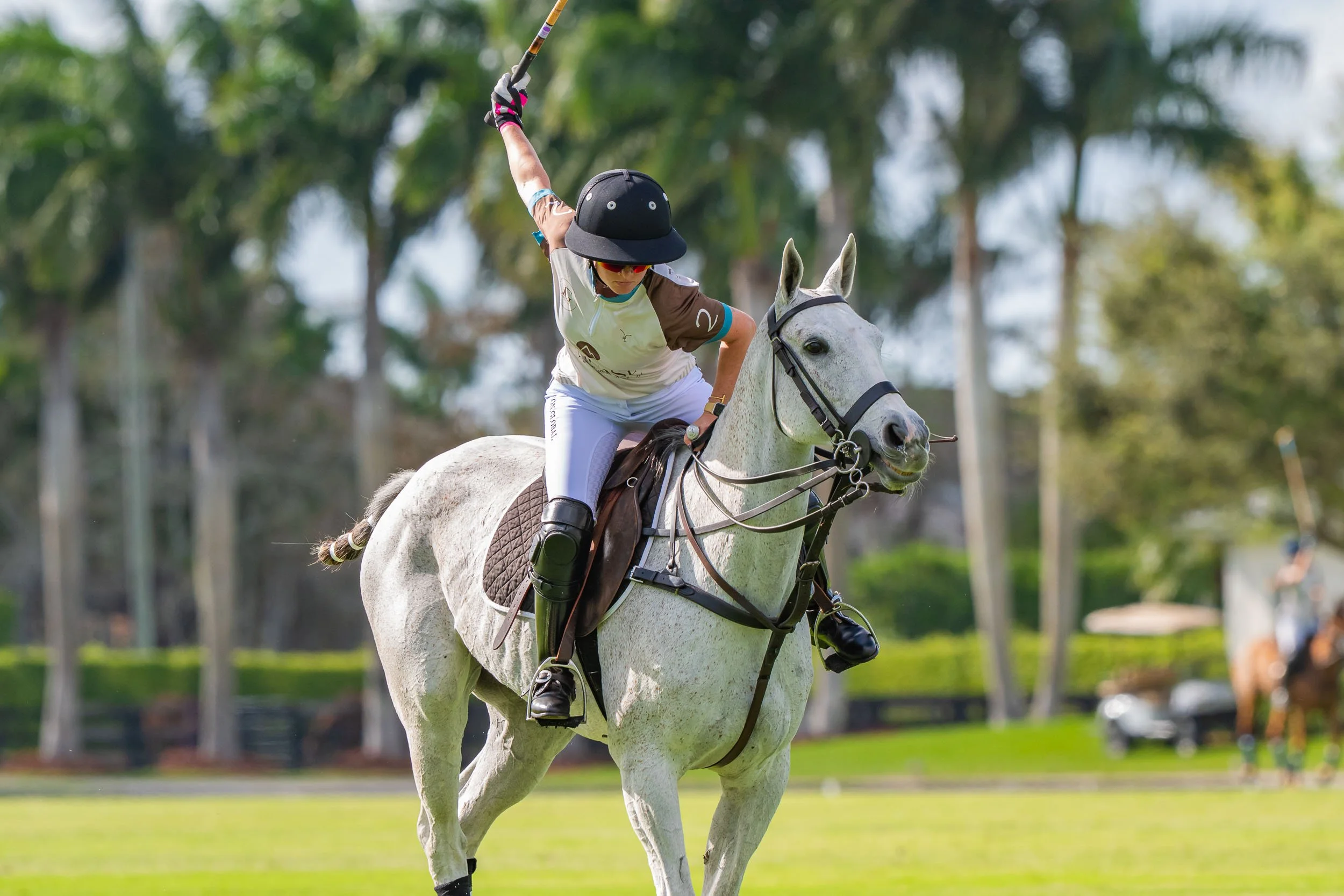 A polo player riding a white horse during a game, wearing a black helmet, sunglasses, and athletic clothing, with trees blurred in the background.