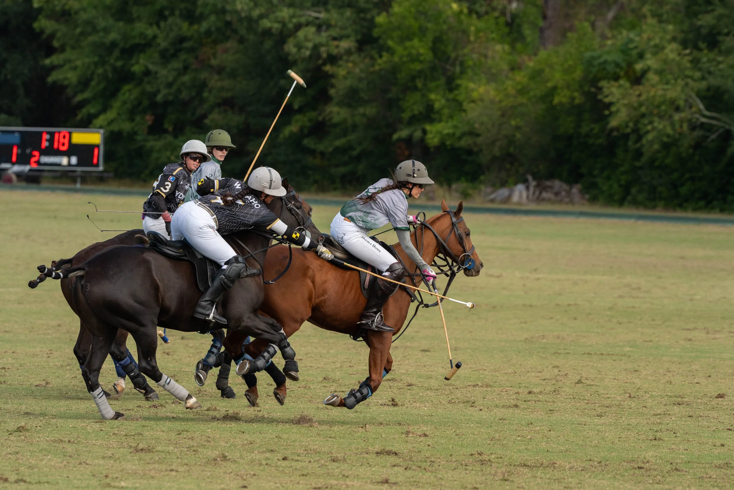 Polo players on horseback competing on a grassy field with a scoreboard in the background